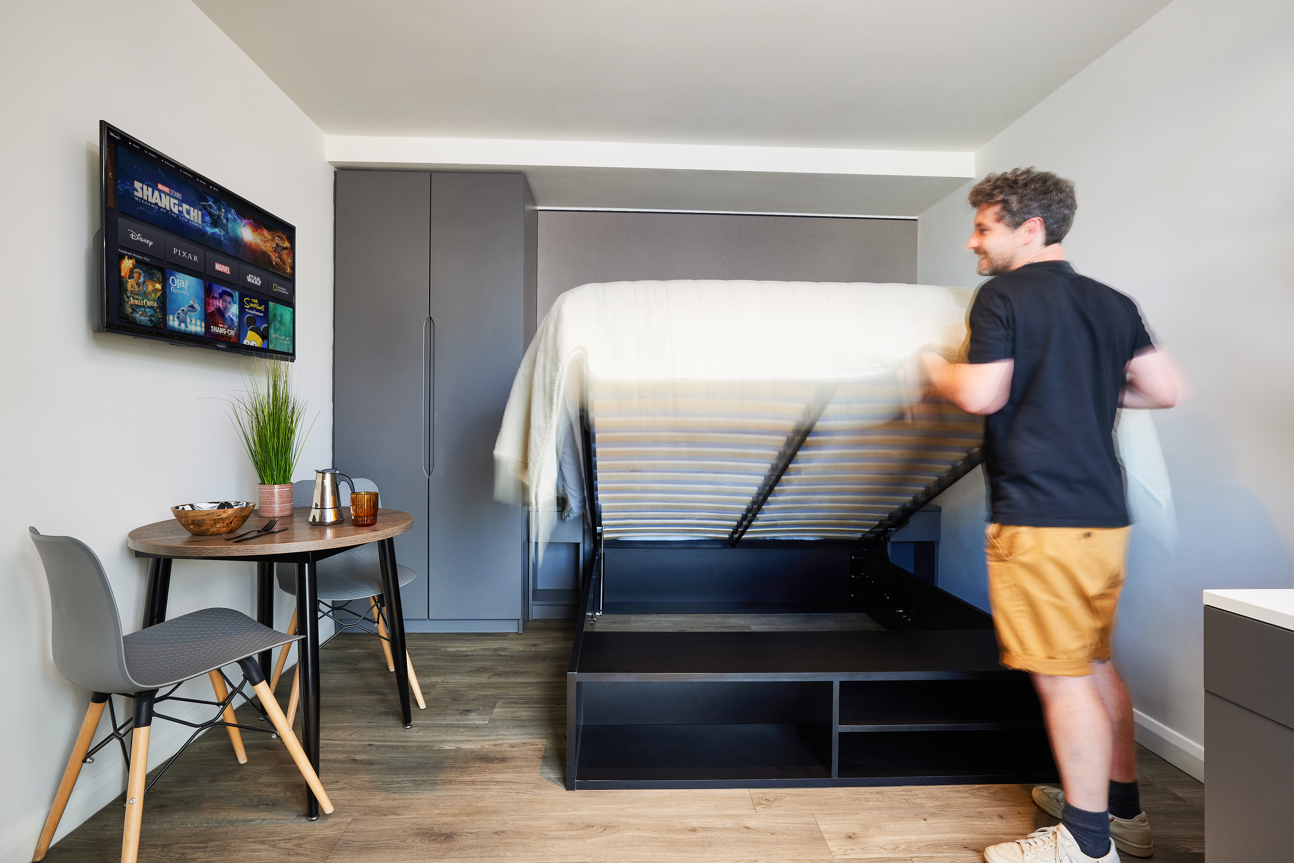 A man in a black shirt and khaki shorts is folding up a wall bed in a small, modern room. The place features a TV on the wall, a small round table with two gray chairs, and a plant. The strawberry wood floor adds warmth to the compact and organized space.