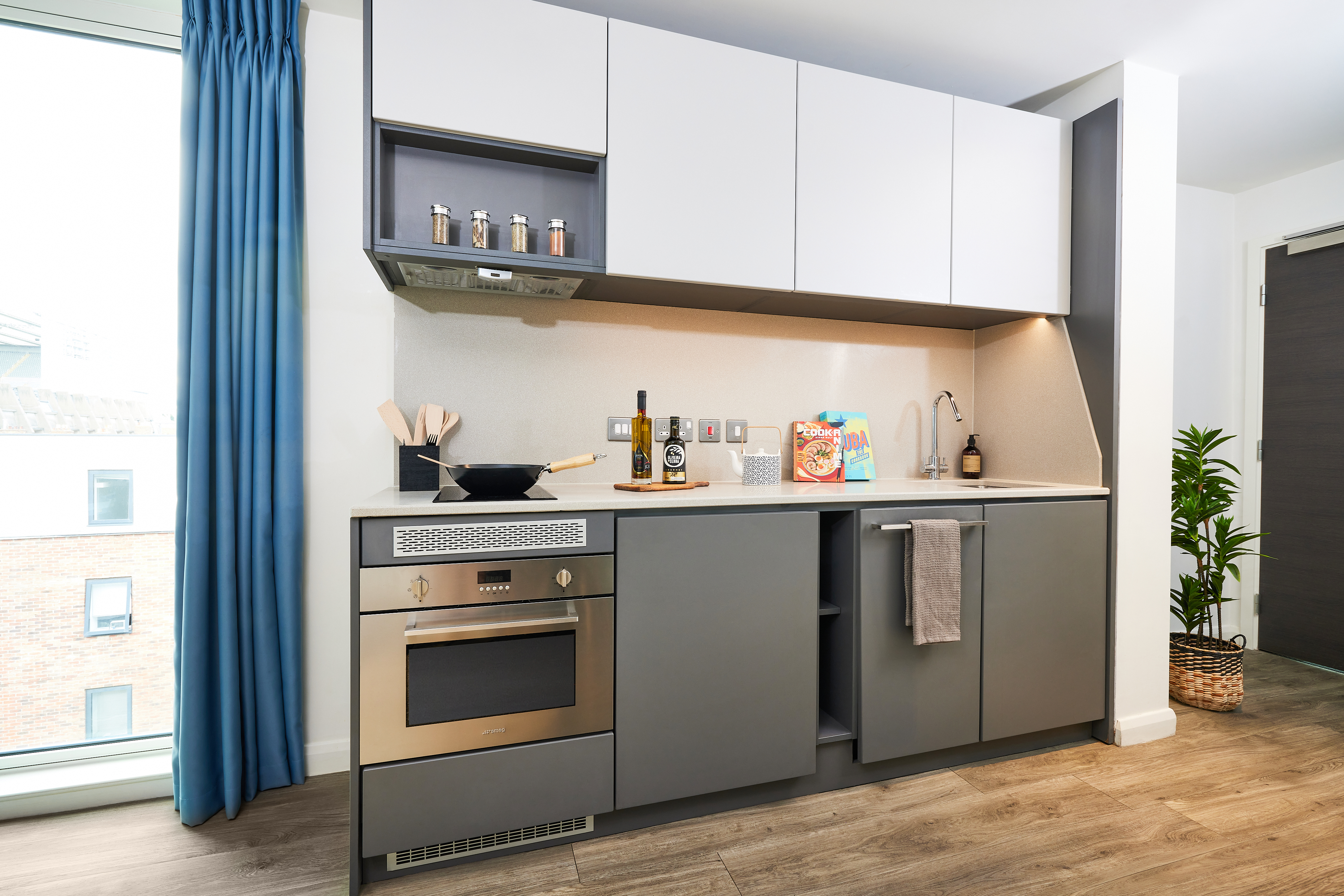 Modern kitchen at Strawberry Place with grey cabinets and white uppers. It features a built-in oven, a sleek countertop with cooking utensils, a bottle, and a cereal box. A window with blue curtains is on the left, and a potted plant is on the floor.