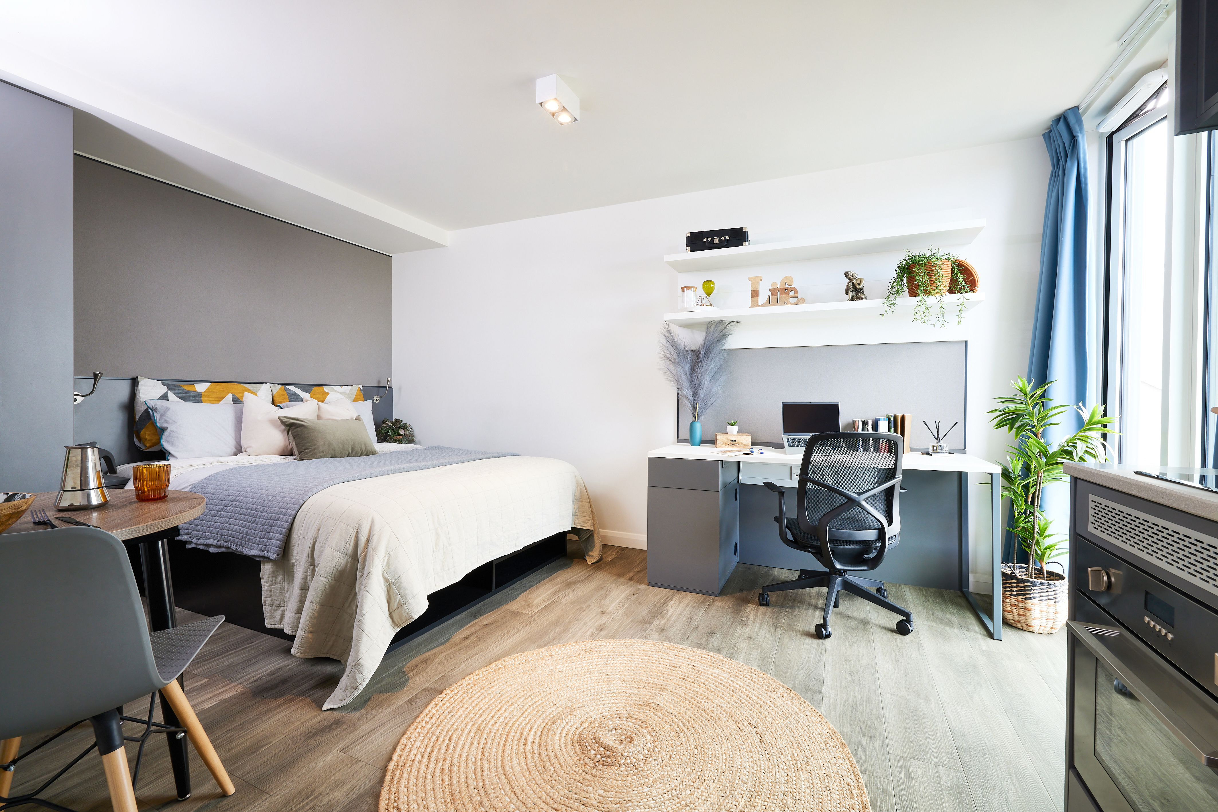 A modern, minimalist bedroom where a bed with white and beige bedding complements a desk with a computer and chair. A round woven rug on wooden flooring adds warmth. Shelves above the desk display plants and decor, infusing the space with charm while large windows drench this tranquil place in natural light.