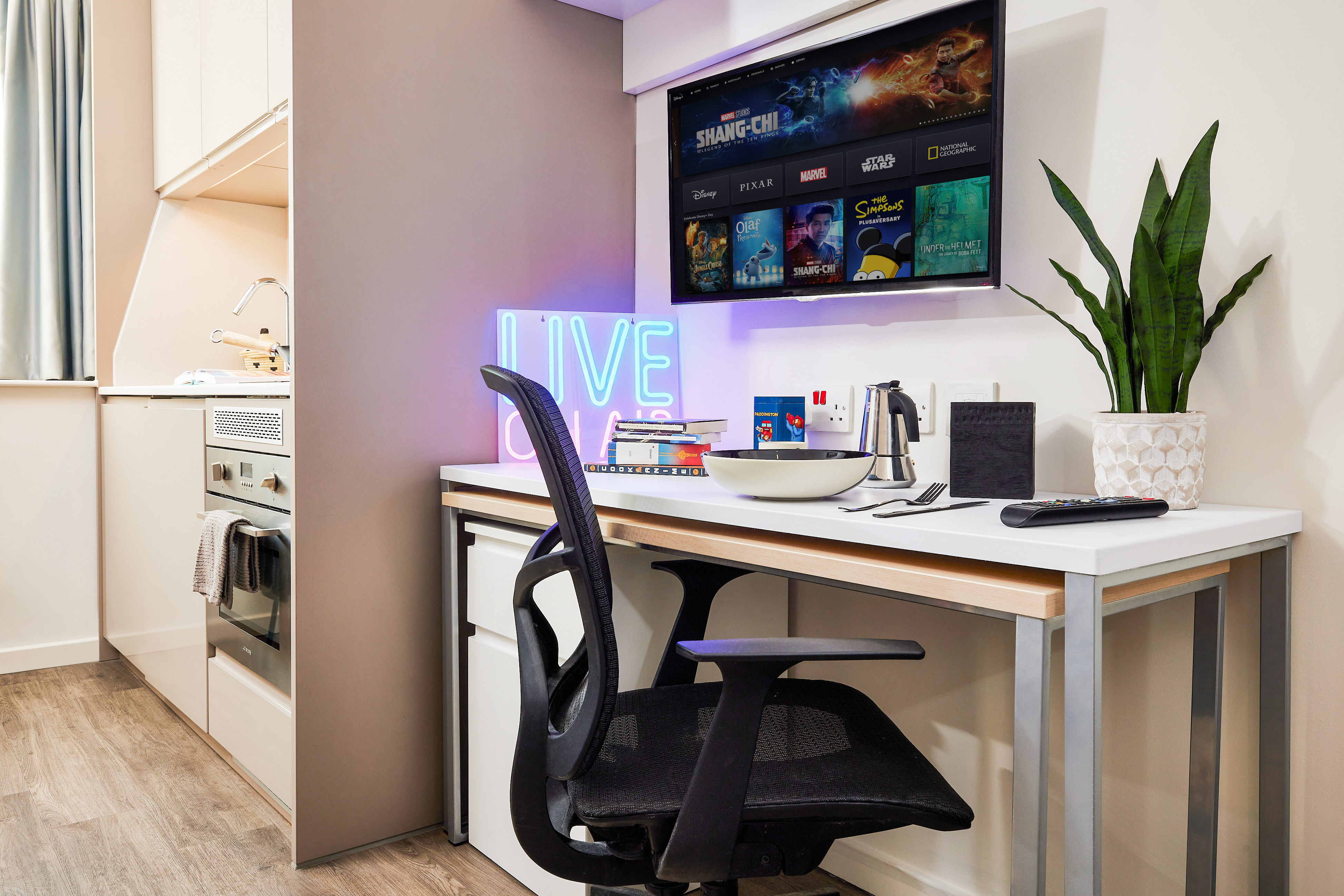 A modern desk setup with a monitor displaying a streaming app, a keyboard, and a pen holder partners perfectly with the glowing "LIVE" neon sign. A bowl sits alongside, while an oven and plant in the background add warmth. Wood flooring and soft lighting complete this harmonious scene.