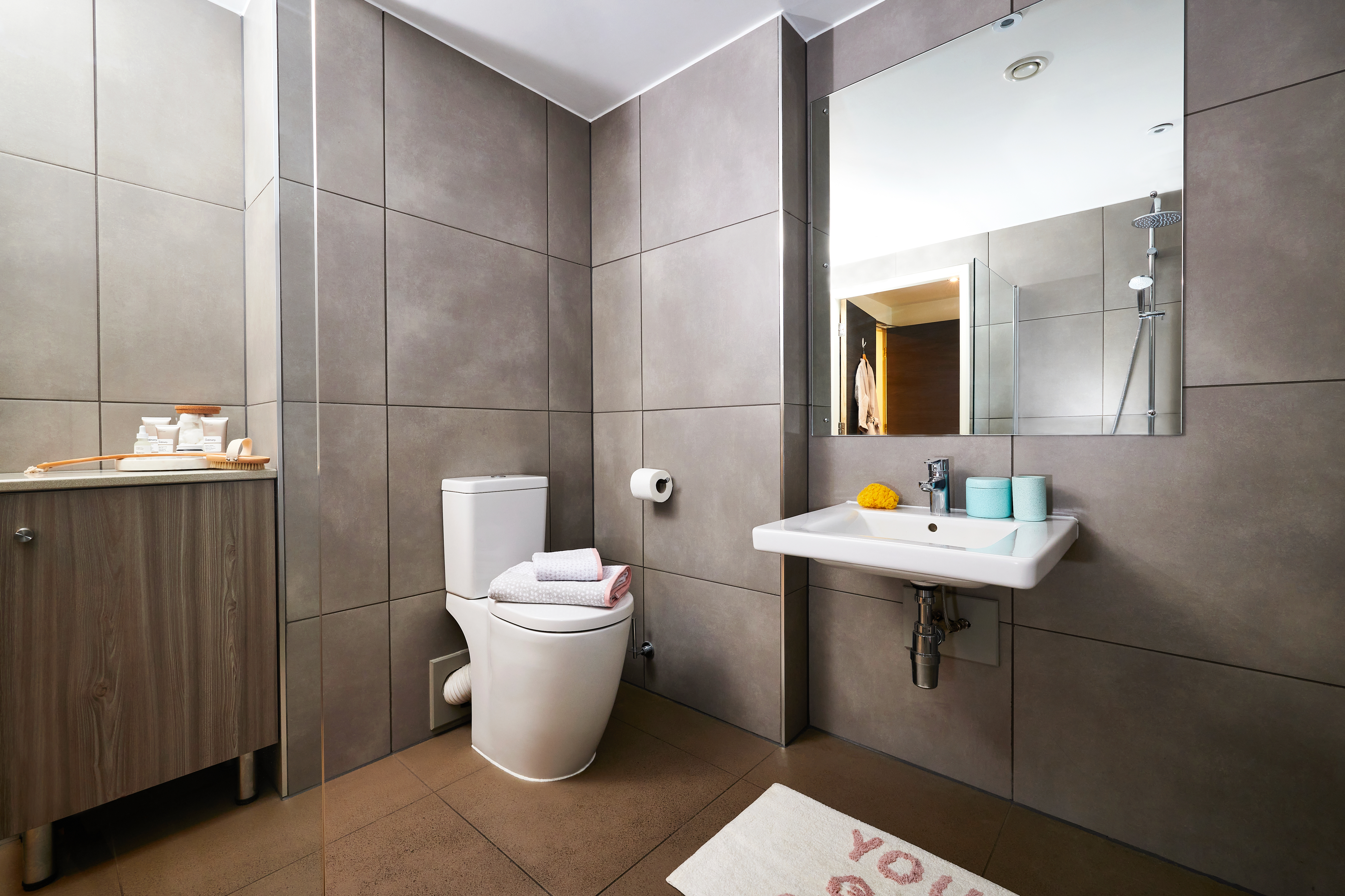 Modern bathroom with gray tiled walls and floors, featuring a square-shaped mirror above the sink. Contains a toilet, a sleek wooden cabinet, and a partially visible shower. Towels and toiletries are neatly arranged on circular shelves, and a decorative mat lies on the floor.