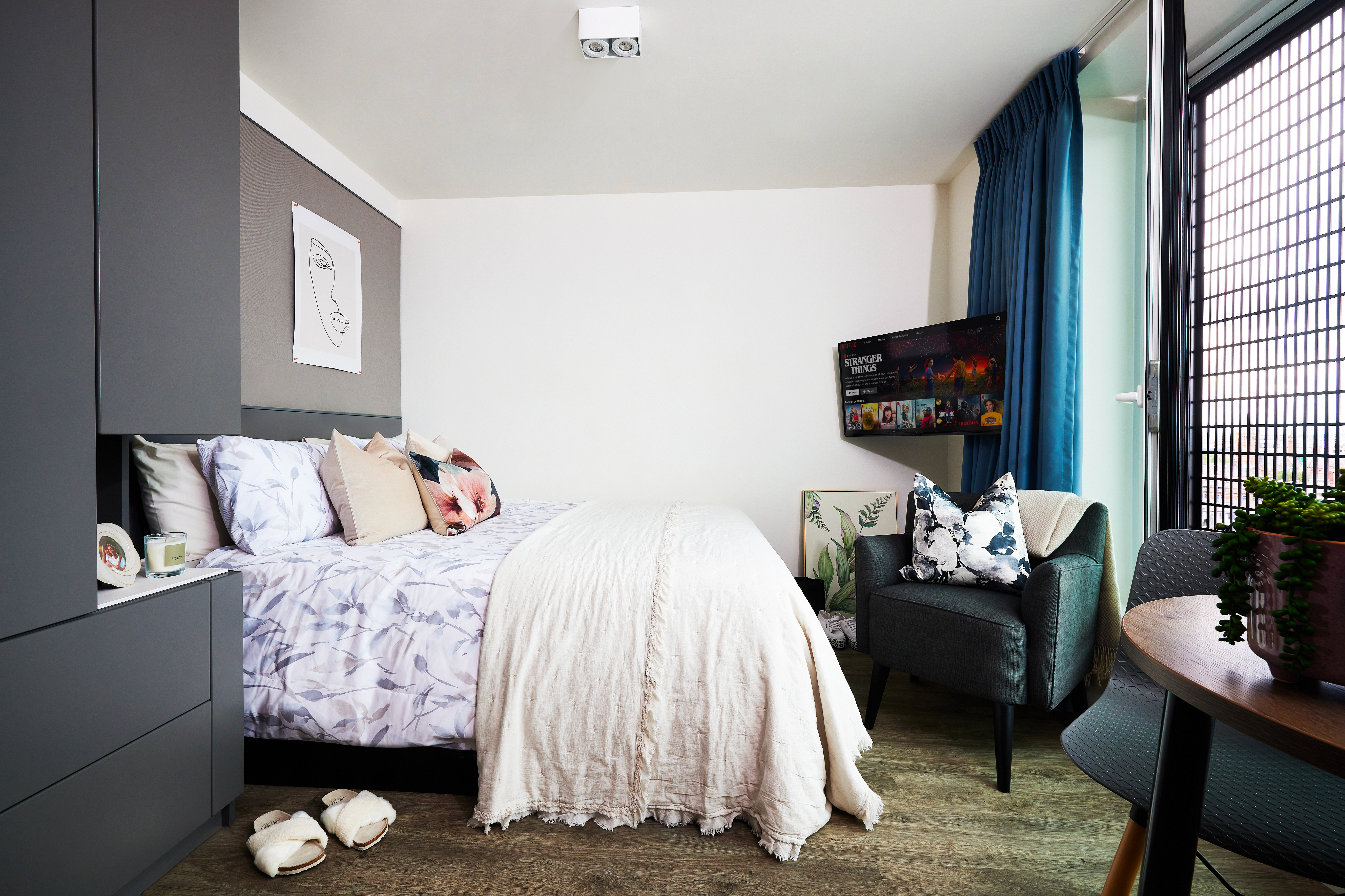 A cozy bedroom features a neatly made bed with floral-patterned pillows and a white comforter. A square TV is mounted on the wall, and a plush chair sits nearby. A large window with blue curtains illuminates the space, which includes a small plant on a round table.