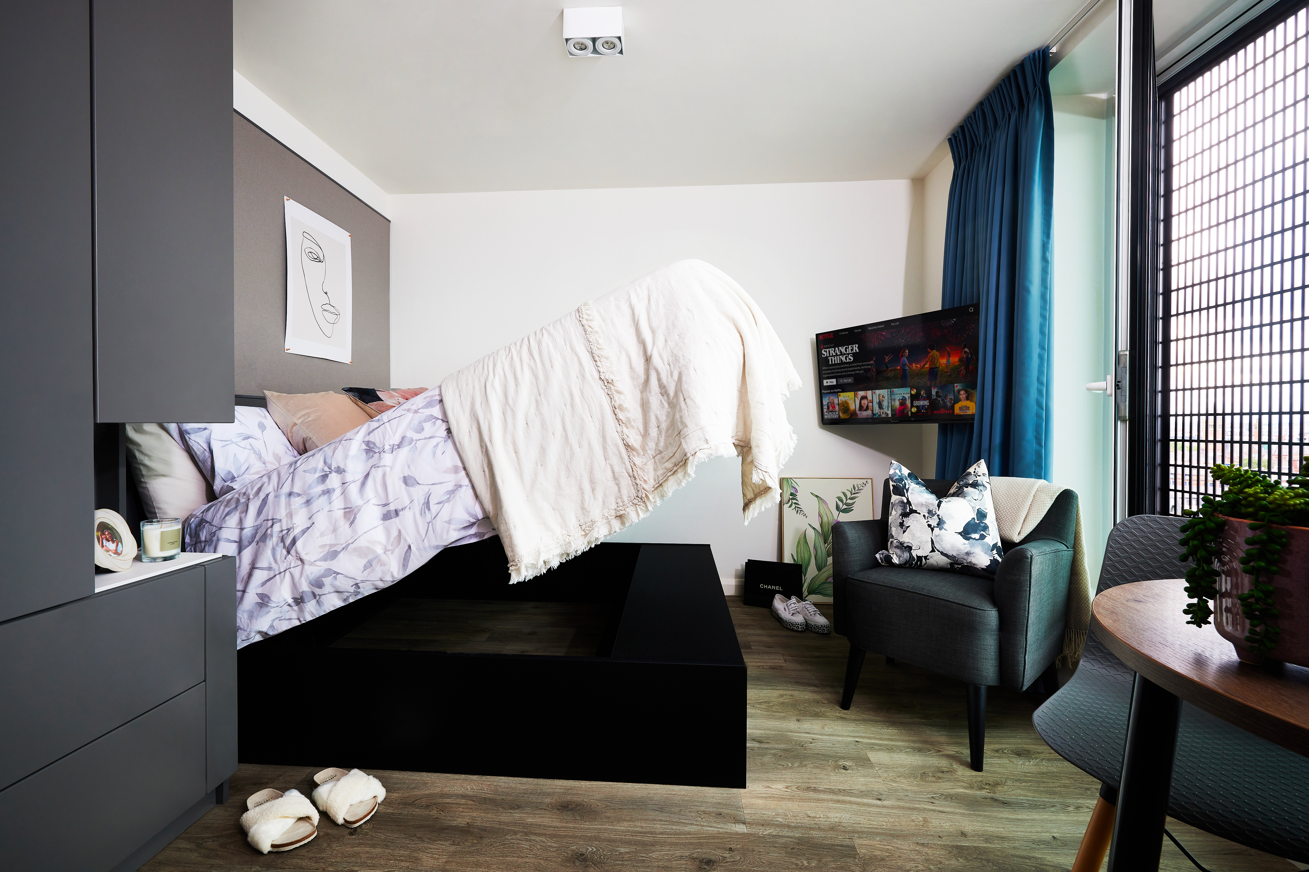 Modern bedroom with a bed lifted to reveal storage underneath. The room features a gray armchair, a wall-mounted TV, light wood flooring, and a large window with blue curtains that hint at the tranquility of Circle Square. Decorative plants and slippers complete the ambiance.