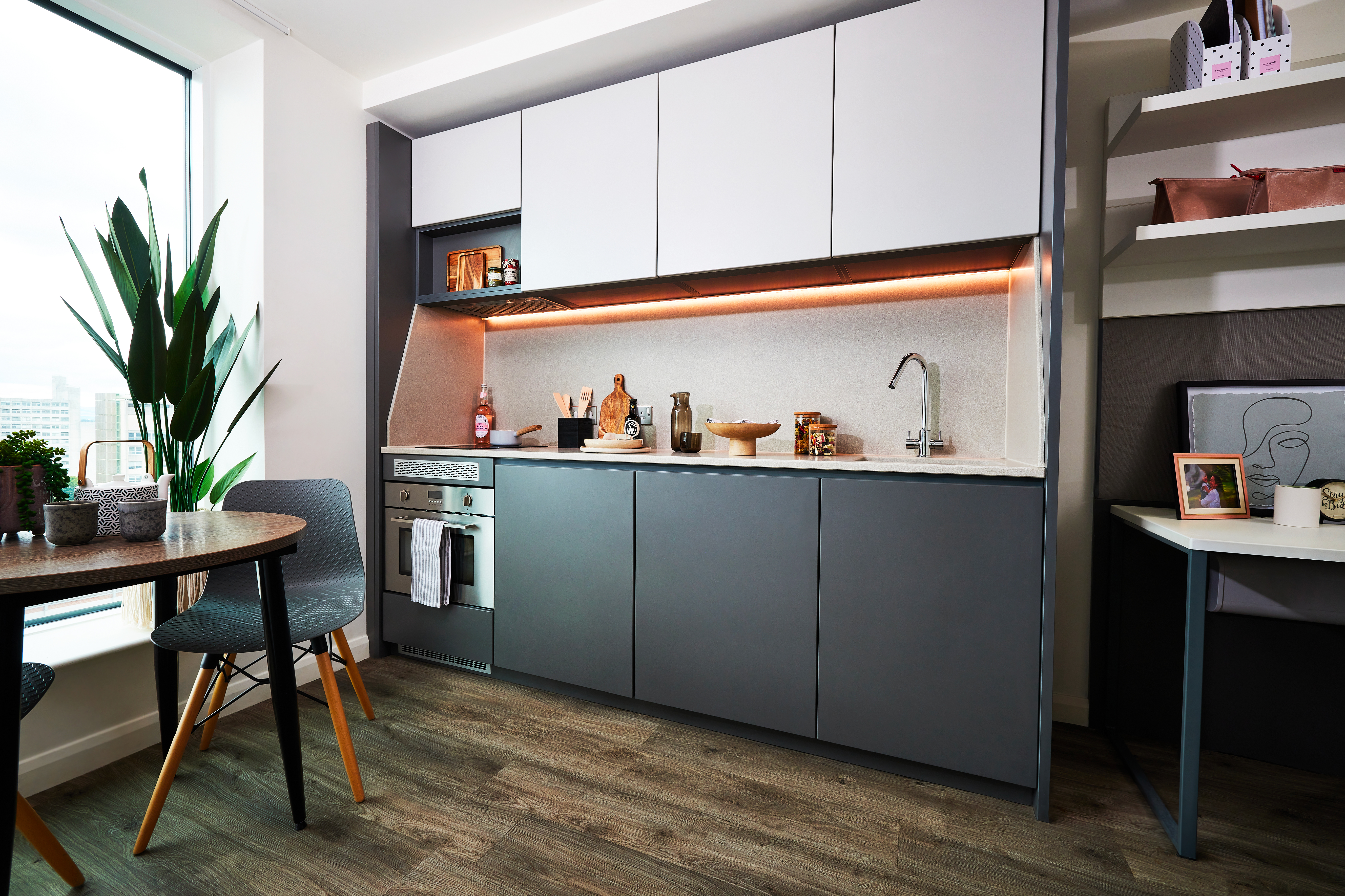 A modern kitchen with sleek gray cabinets, an oven, and a small stovetop. There's under-cabinet lighting highlighting the backsplash. A small round table with chairs forms a cozy circle by the window, complemented by a large potted plant. Warm wooden flooring completes the scene.