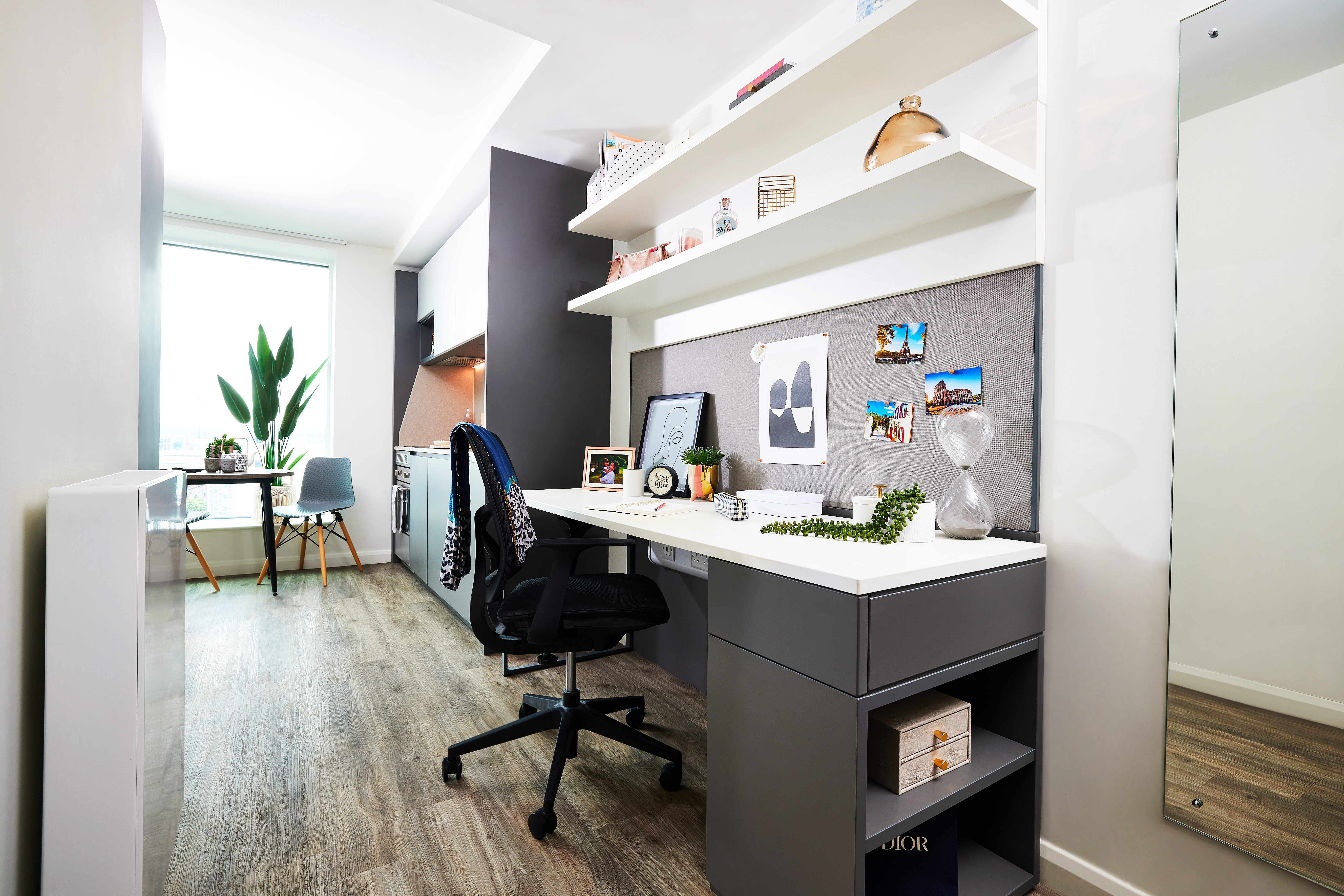 A modern home office with a white desk and black chair is complemented by square shelves holding books and decor above. A corkboard displays photos, while a large circular mirror graces the right side. The window hosts a potted plant and chair in the background, set against wood flooring throughout.