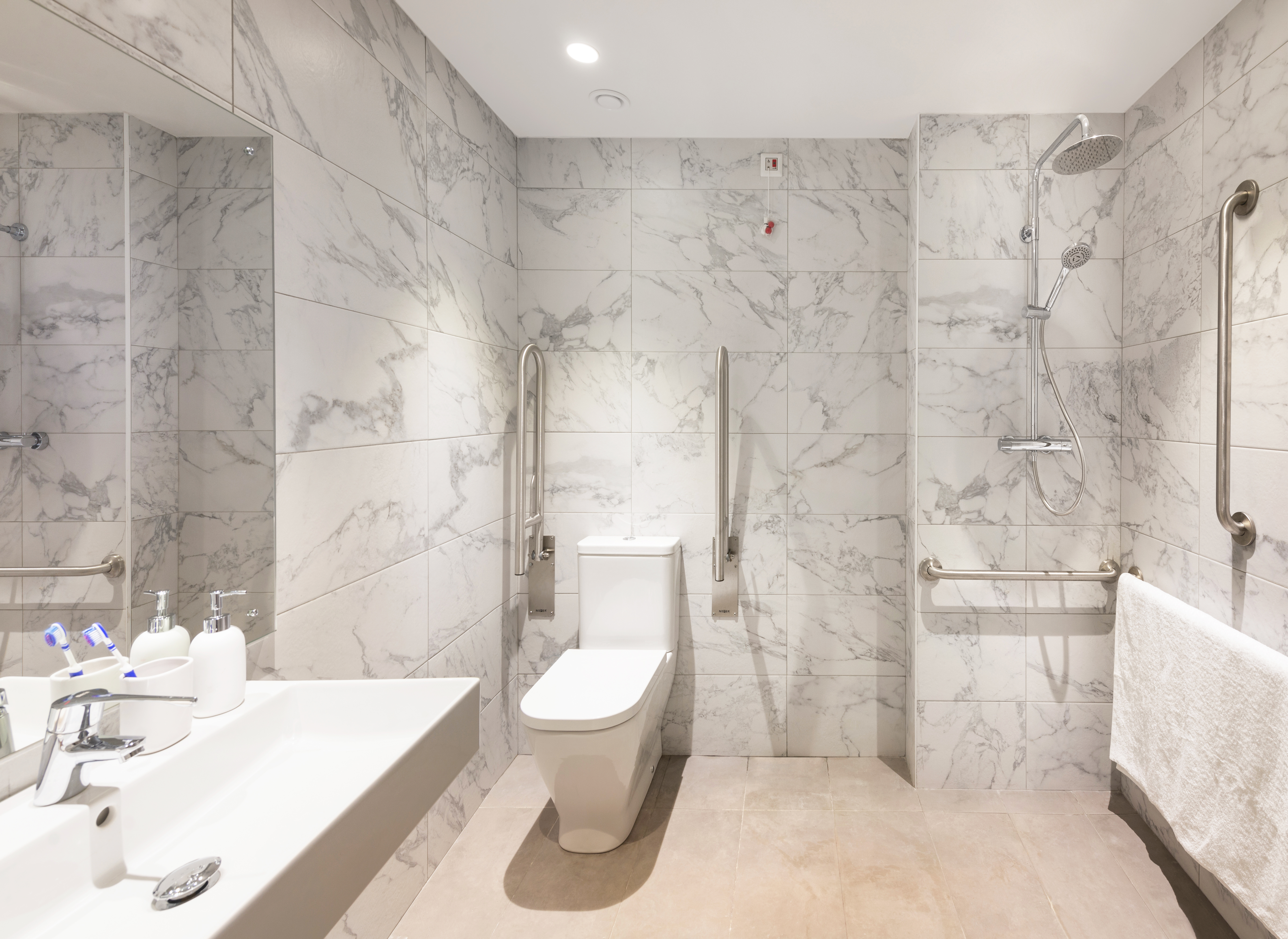 Spacious modern bathroom with white marble walls, featuring a walk-in shower with overhead and hand-held showerheads, a white toilet, and a wall-mounted sink. There are multiple grab bars, a towel rack, and a mirror above the sink.