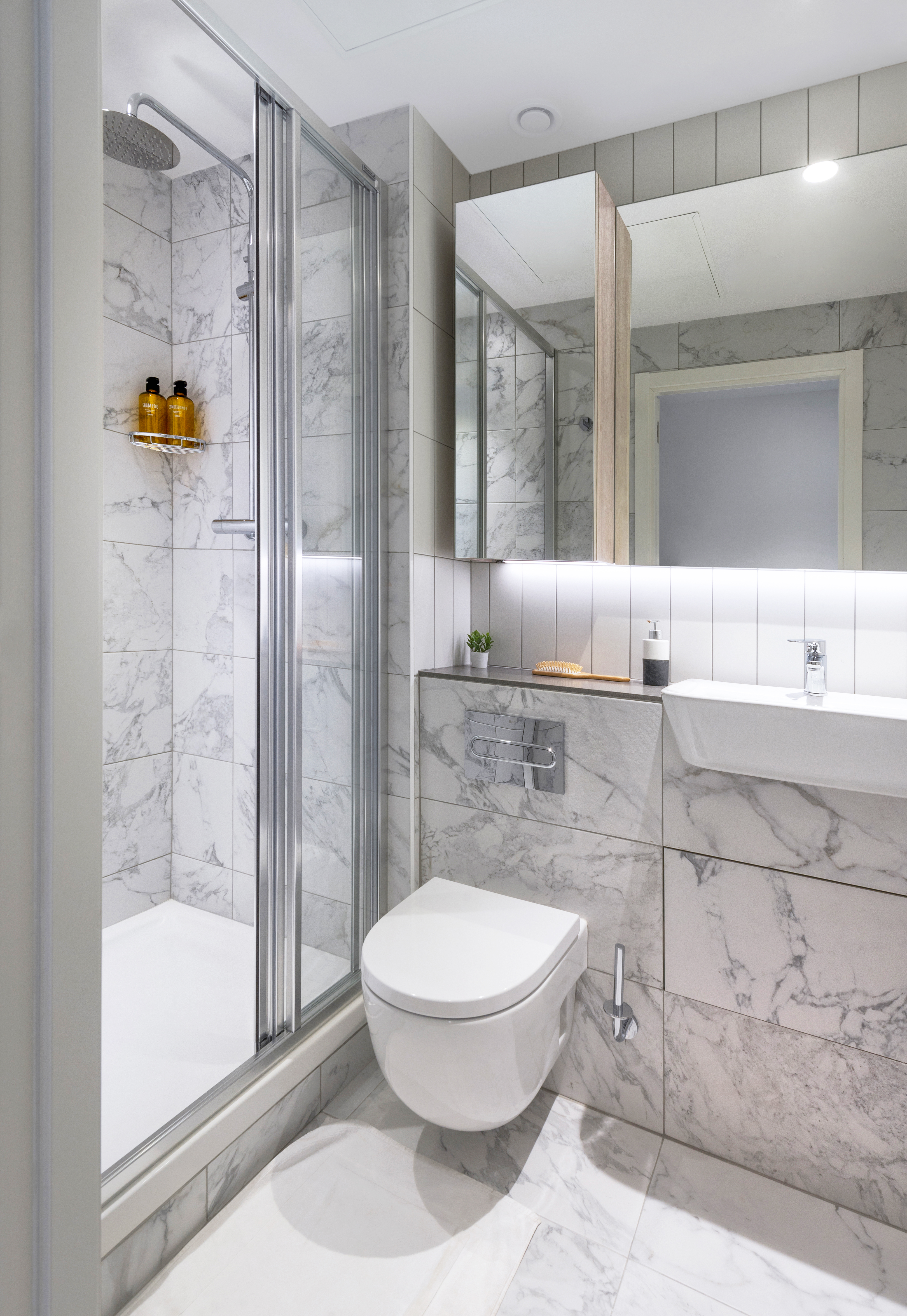 Modern bathroom with marble tiles, a glass-enclosed shower, and a wall-mounted toilet. The white sink features a sleek design with a small plant and soap dispenser on the counter. A large mirror reflects the space, enhancing its brightness.