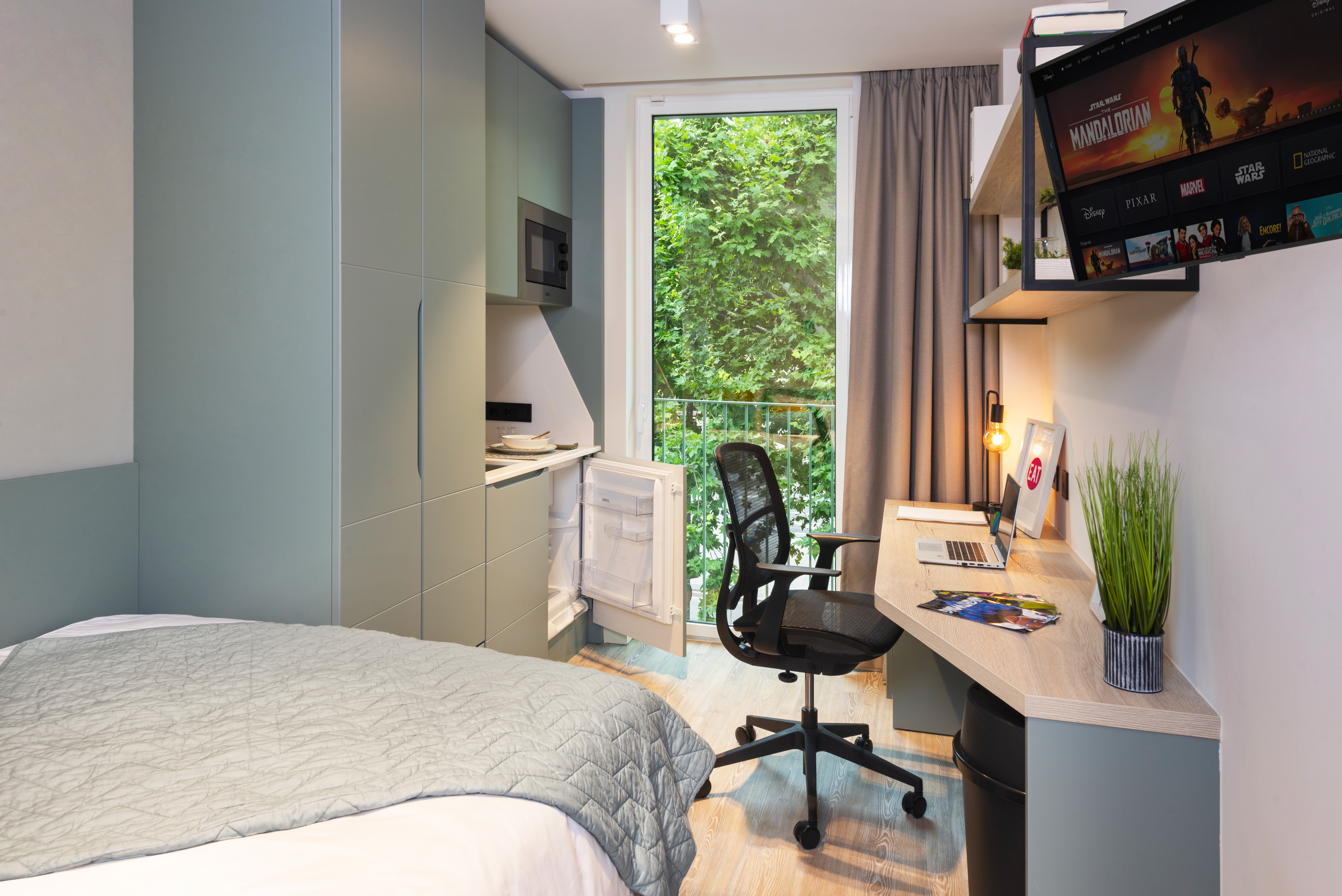Modern dorm room with a bed, desk, and chair. A laptop and plant sit on the desk, and a wall-mounted TV shows a streaming app. Cabinets and a small fridge are on one side, and a large window with leafy view is opposite.