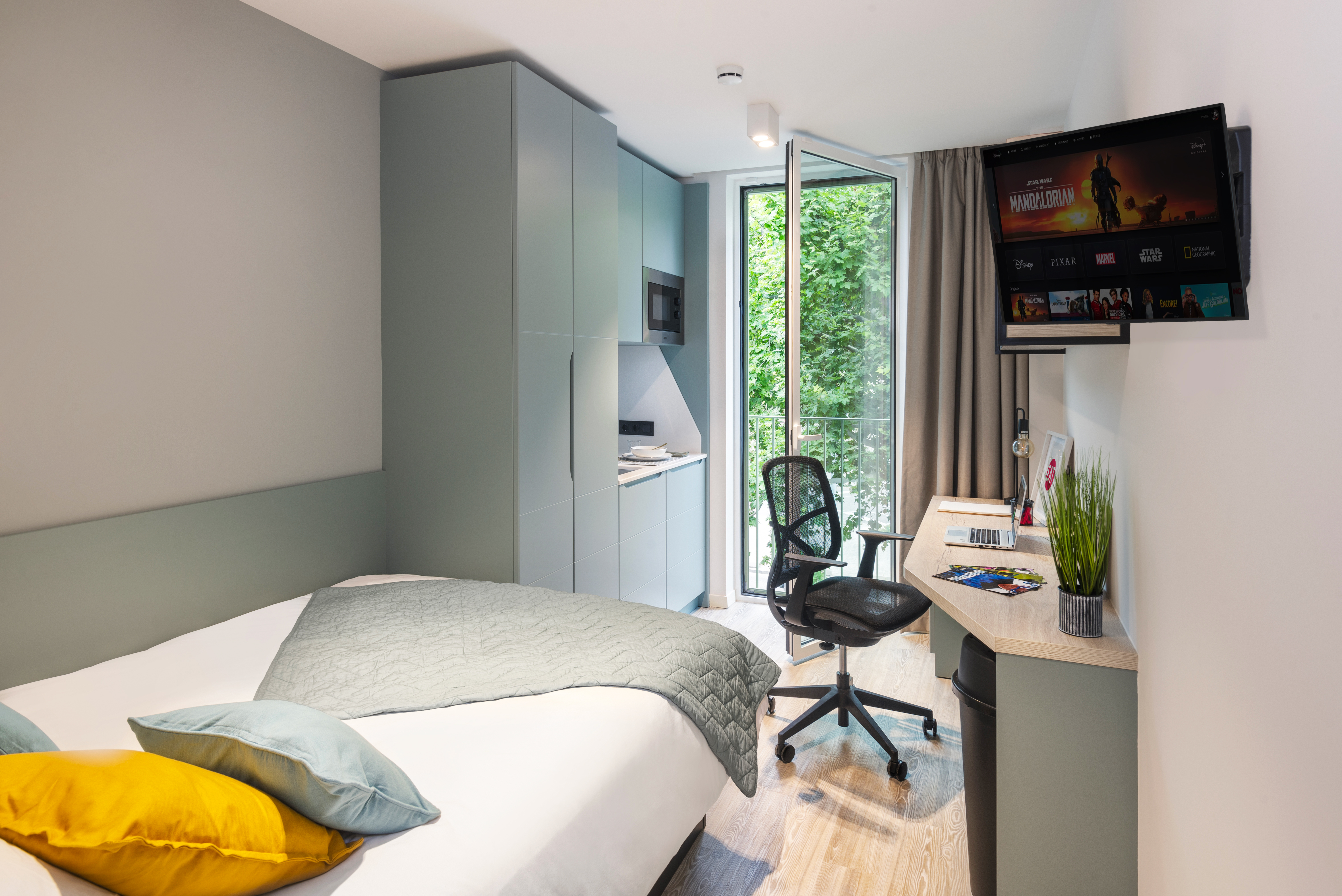 Modern hotel room with a large bed, green bedspread, and pillows. A workspace with a chair, desk, and laptop is next to a window overlooking greenery. Above the desk, a TV is mounted on the wall. The room has light-colored walls and wood flooring.
