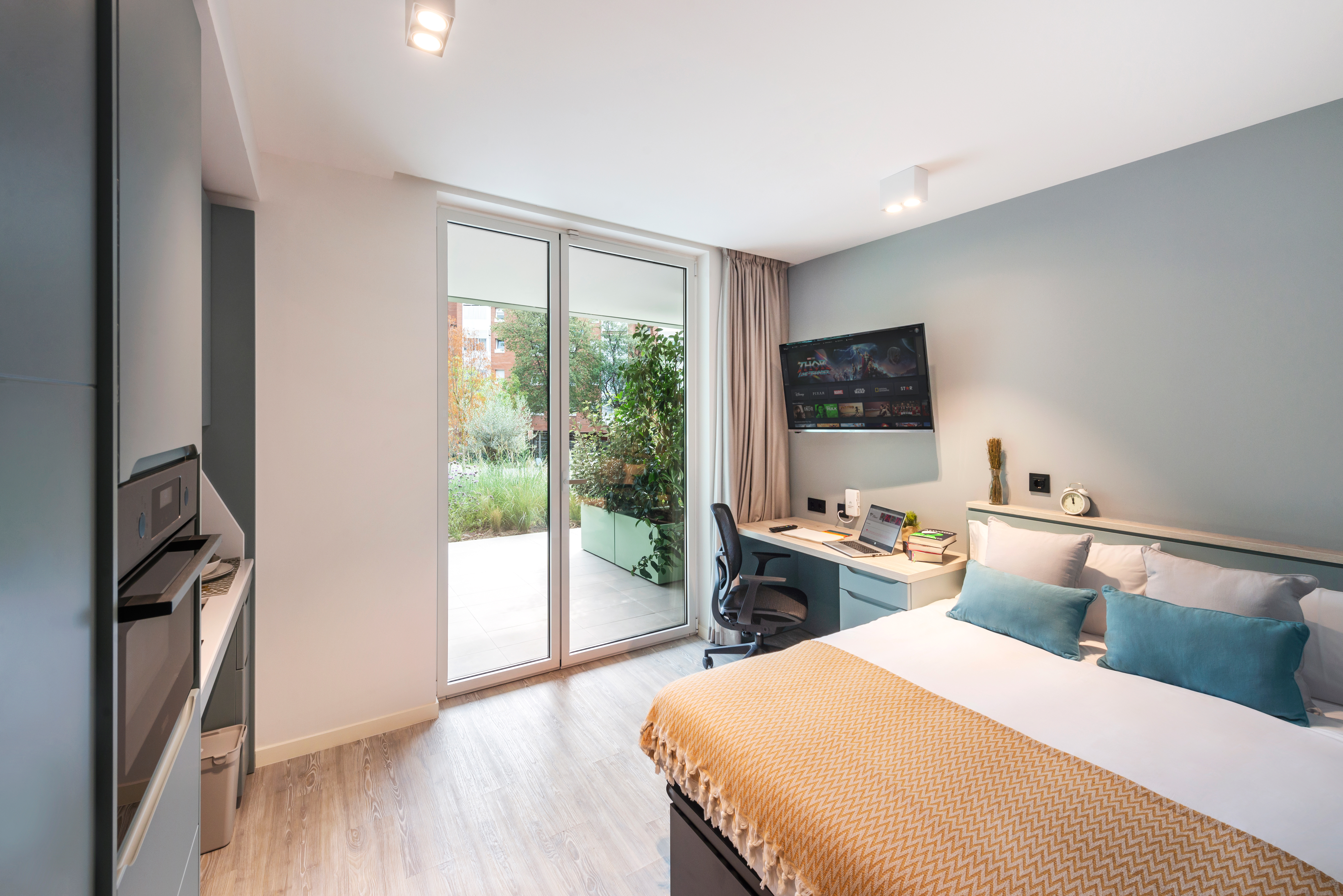 Modern bedroom with a large bed, blue accent pillows, and a yellow throw blanket. A desk with a chair and computer is near floor-to-ceiling windows leading to a garden view. A wall-mounted TV is above the desk. Light wood flooring complements the neutral decor.