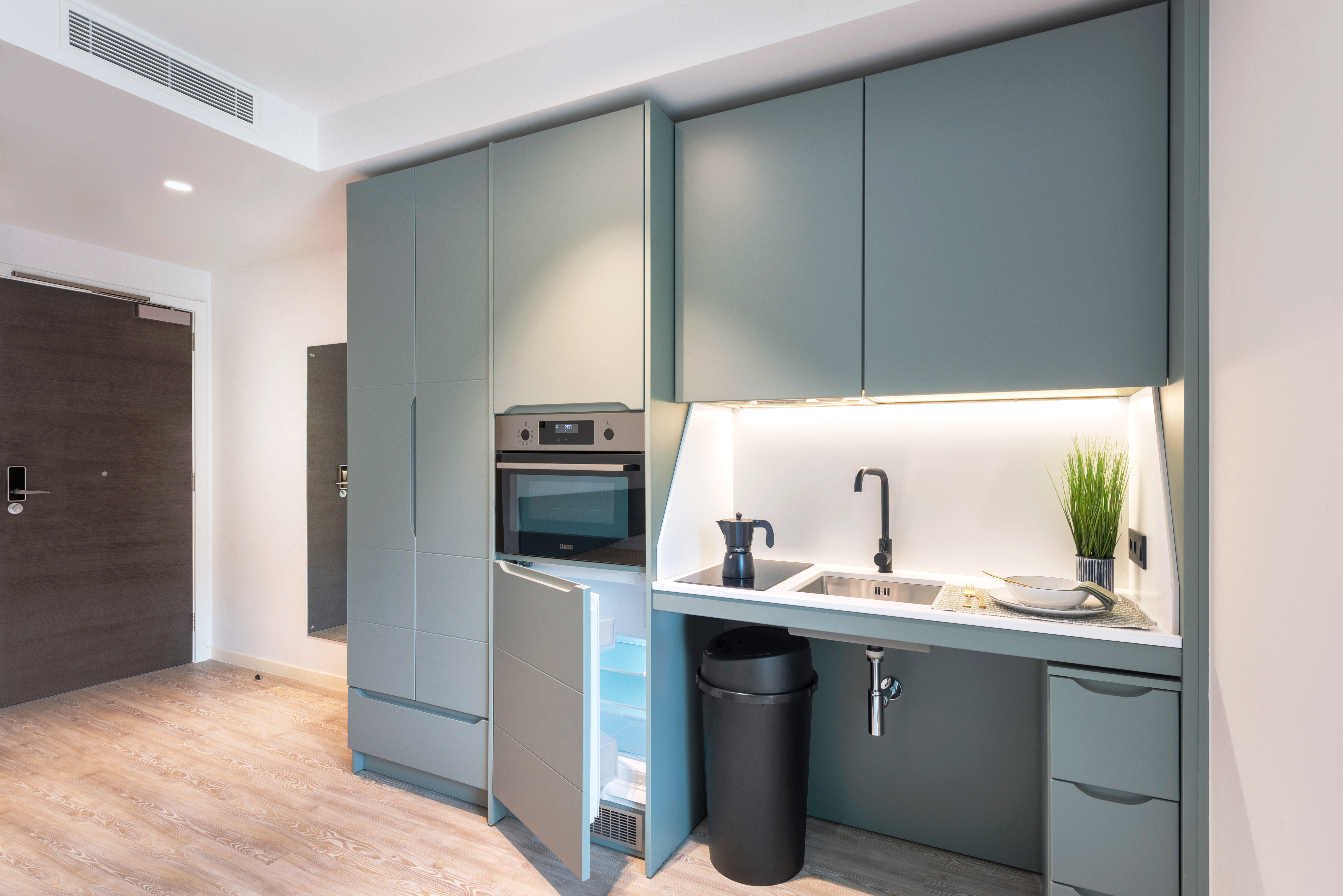Compact modern kitchen with gray cabinets, integrated oven, and fridge with door ajar. A sink with a black faucet sits under bright under-cabinet lighting next to a small potted plant and dishes. Light wood flooring and a dark wooden door are visible.