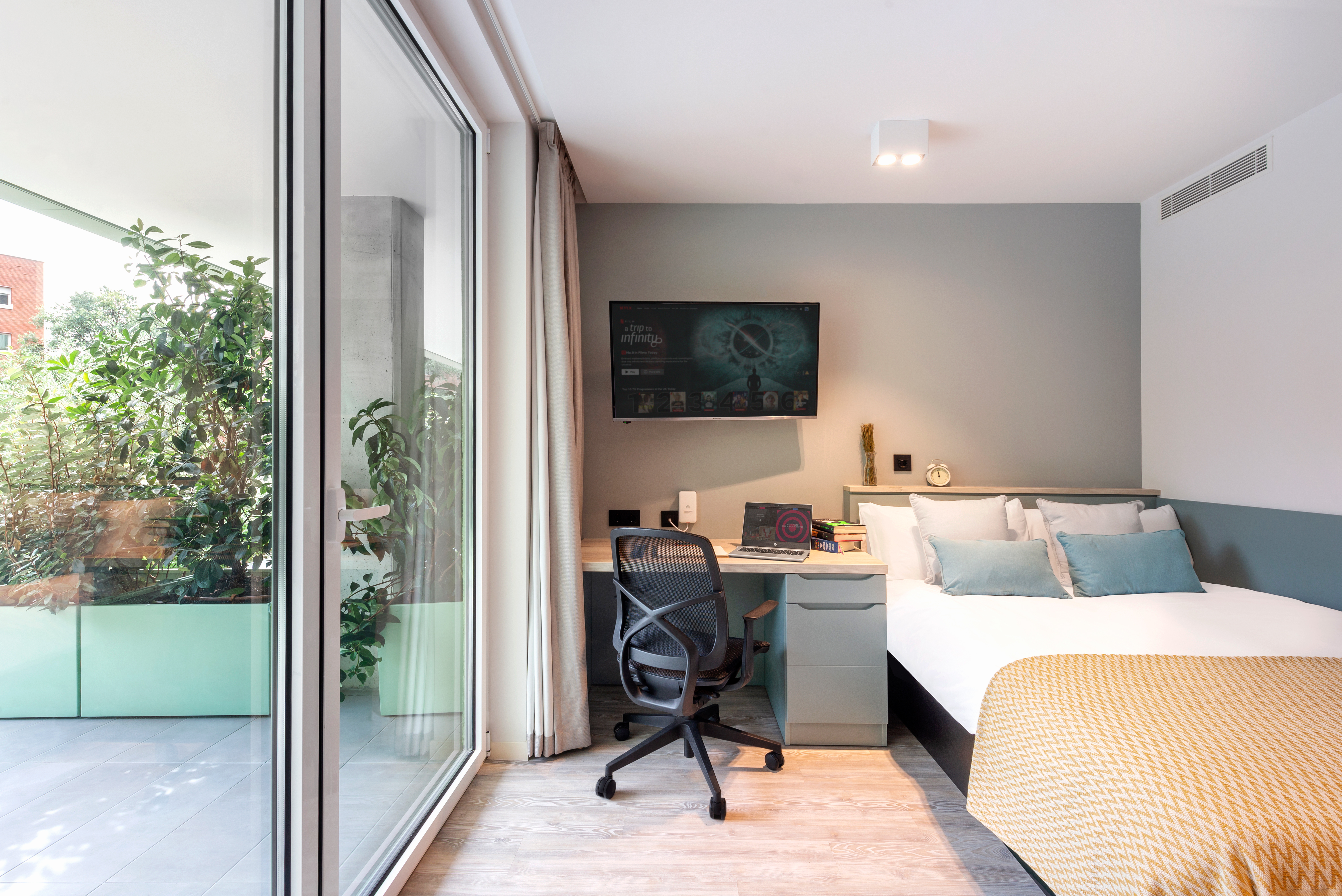 A modern bedroom with a large glass door opening to a balcony with plants. Inside, there's a bed with white and mustard bedding, a desk with a chair, a wall-mounted TV, and a light gray and teal color scheme. Natural light fills the room.