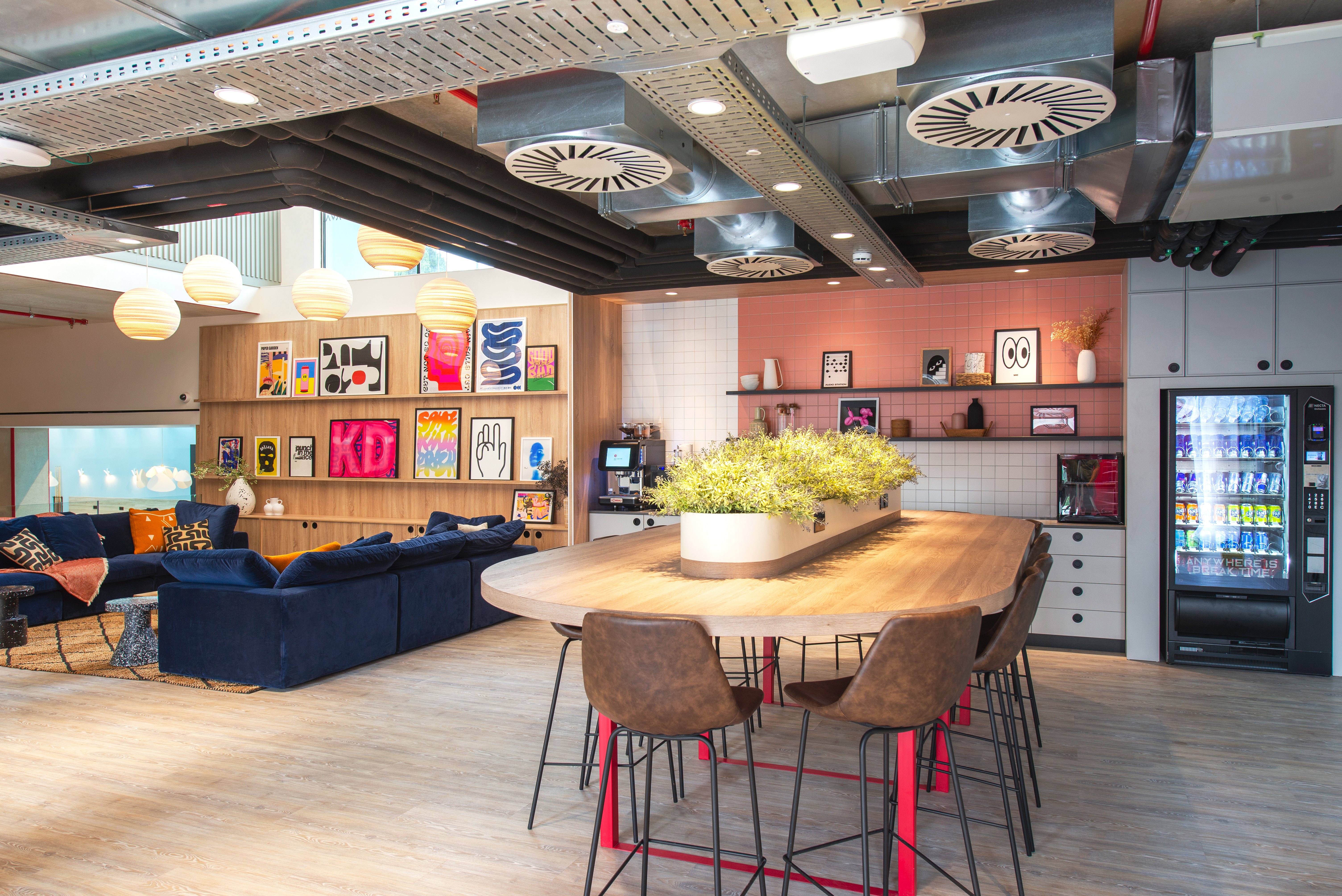 A modern, vibrant office lounge with a large wooden table surrounded by bar stools. The room features a blue sofa, colorful wall art, pendant lights, and a vending machine. Exposed ducts add an industrial touch to the ceiling.