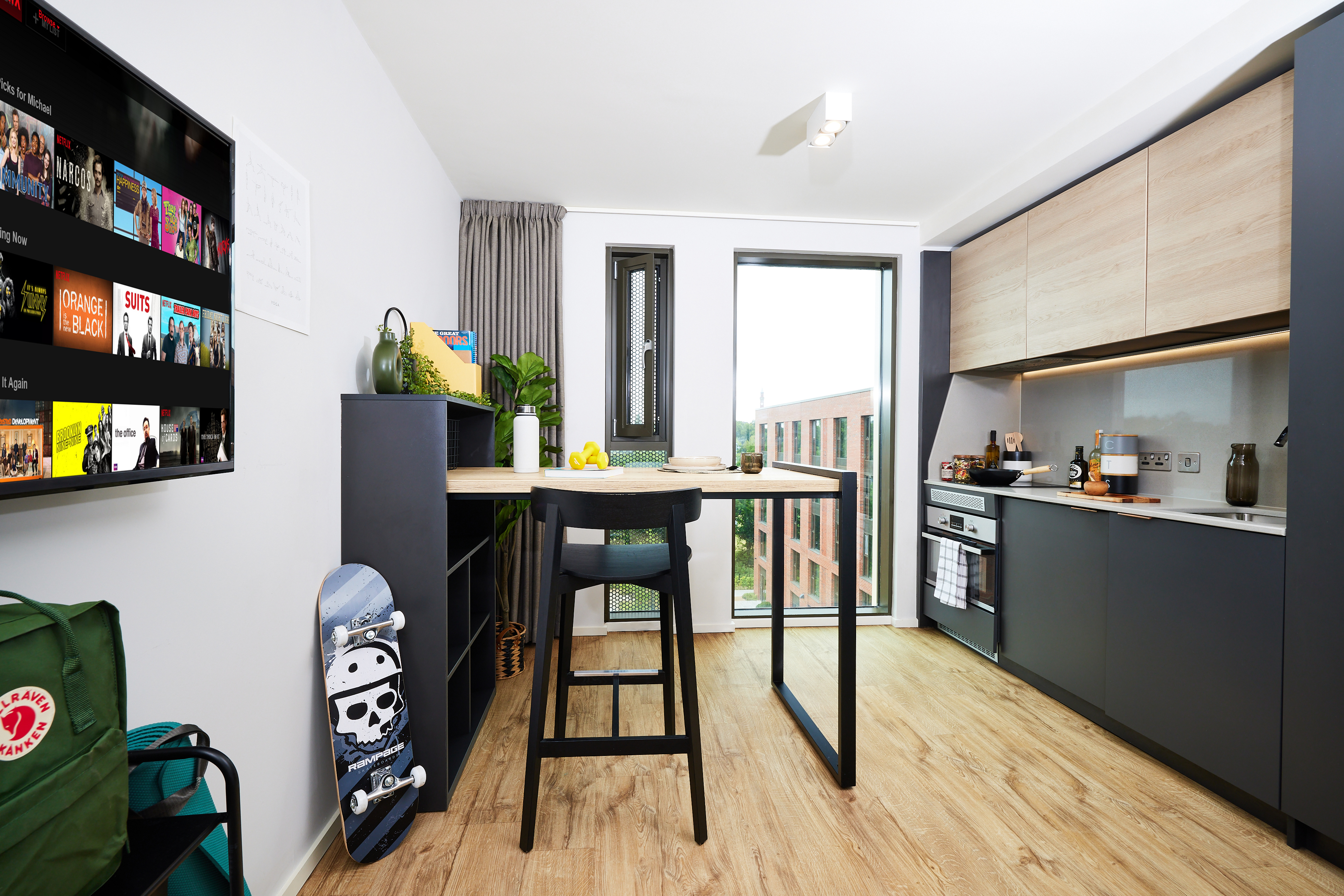 Modern kitchen with sleek cabinets, large window, and wooden flooring. A high table with stools is in the center. A skateboard and a TV with streaming apps are visible. The room is bright and tidy, with some plants adding a touch of greenery.