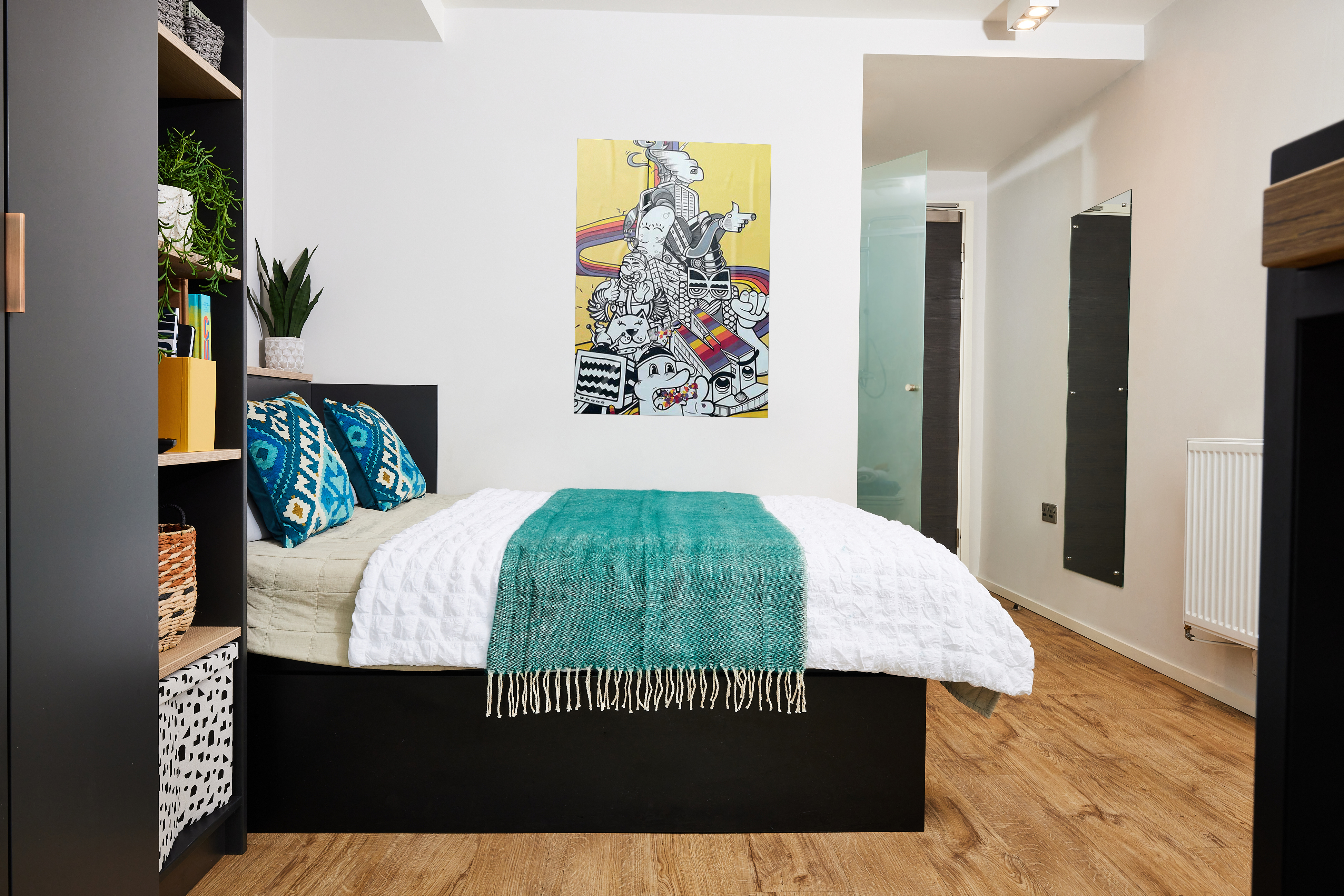 A modern bedroom featuring a single bed with a white quilt and teal blanket. Two blue-patterned pillows are on the bed. A colorful abstract artwork hangs on the wall. Wooden flooring and shelves with plants and baskets decorate the space.