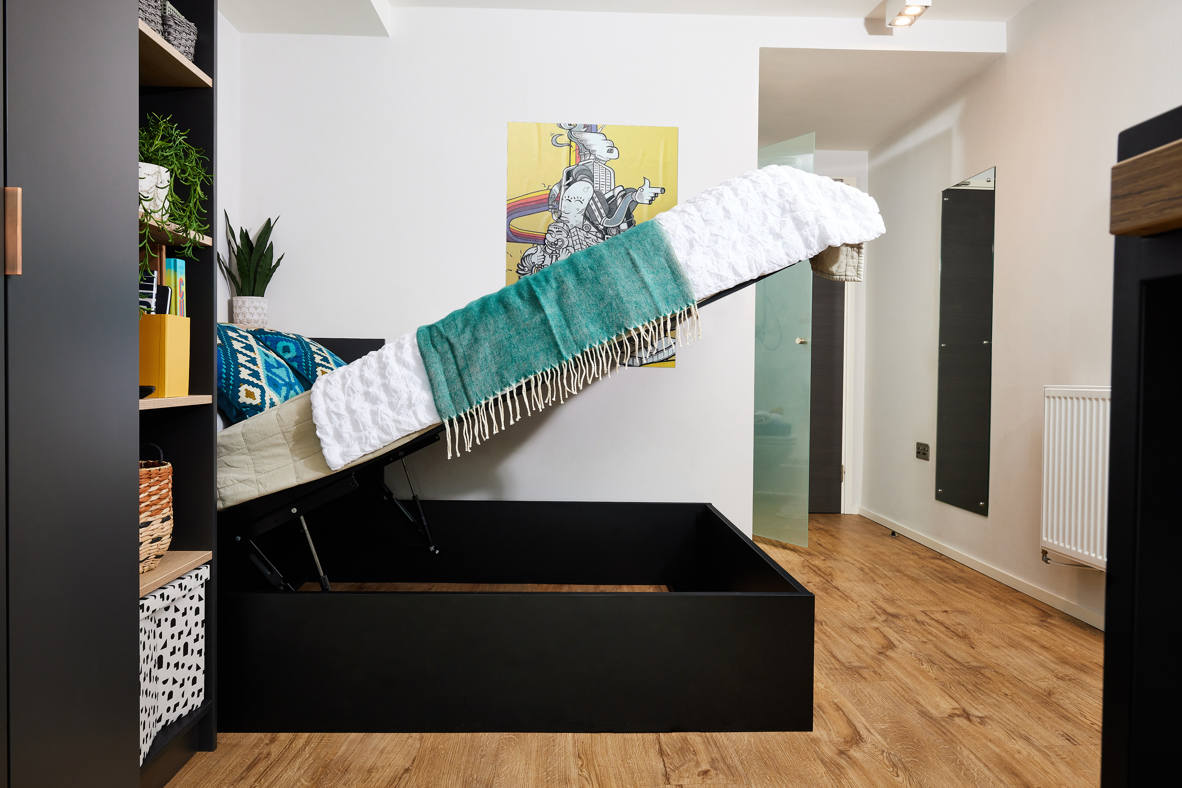 A modern room with a wooden floor features a black storage bed with the mattress uplifted. The wall displays a colorful abstract painting. A tall open shelf on the left contains baskets and plants, adding a decorative touch to the minimalist space.