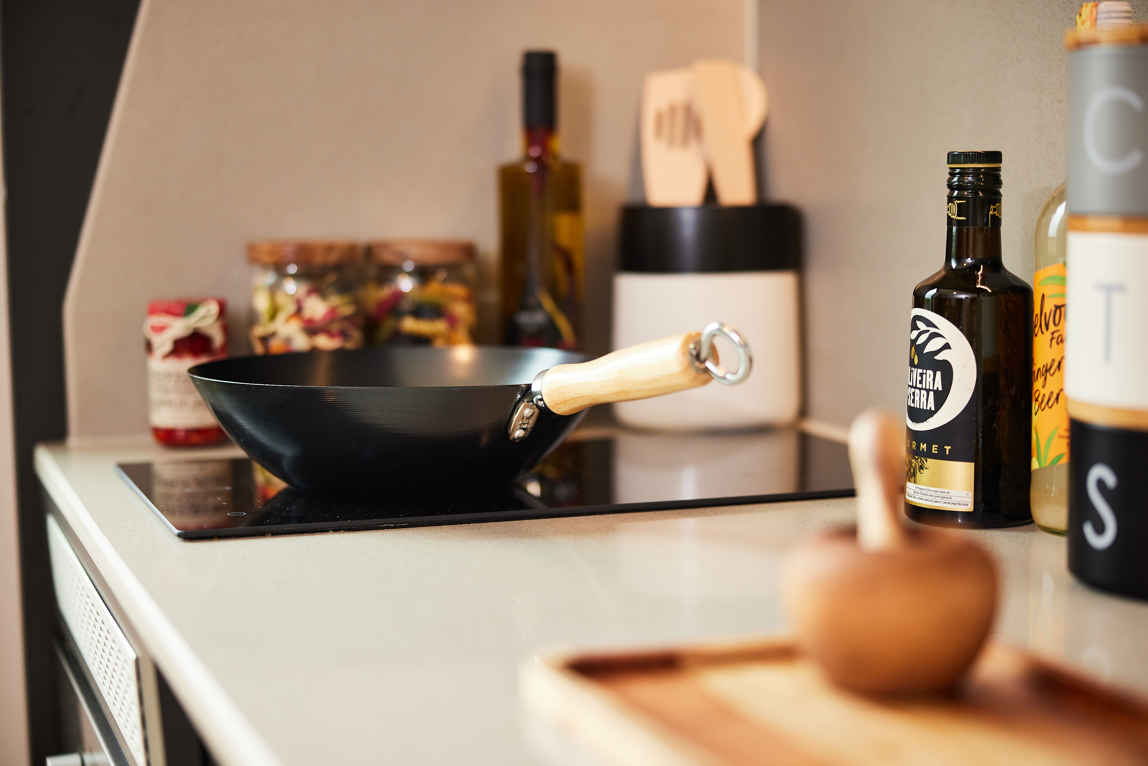 A Portland House kitchen showcases a stovetop with a black pan featuring a wooden handle. Nearby, you'll find various bottles, a jar of preserved food, and a container with utensils alongside a wooden mortar and pestle resting on the cutting board.