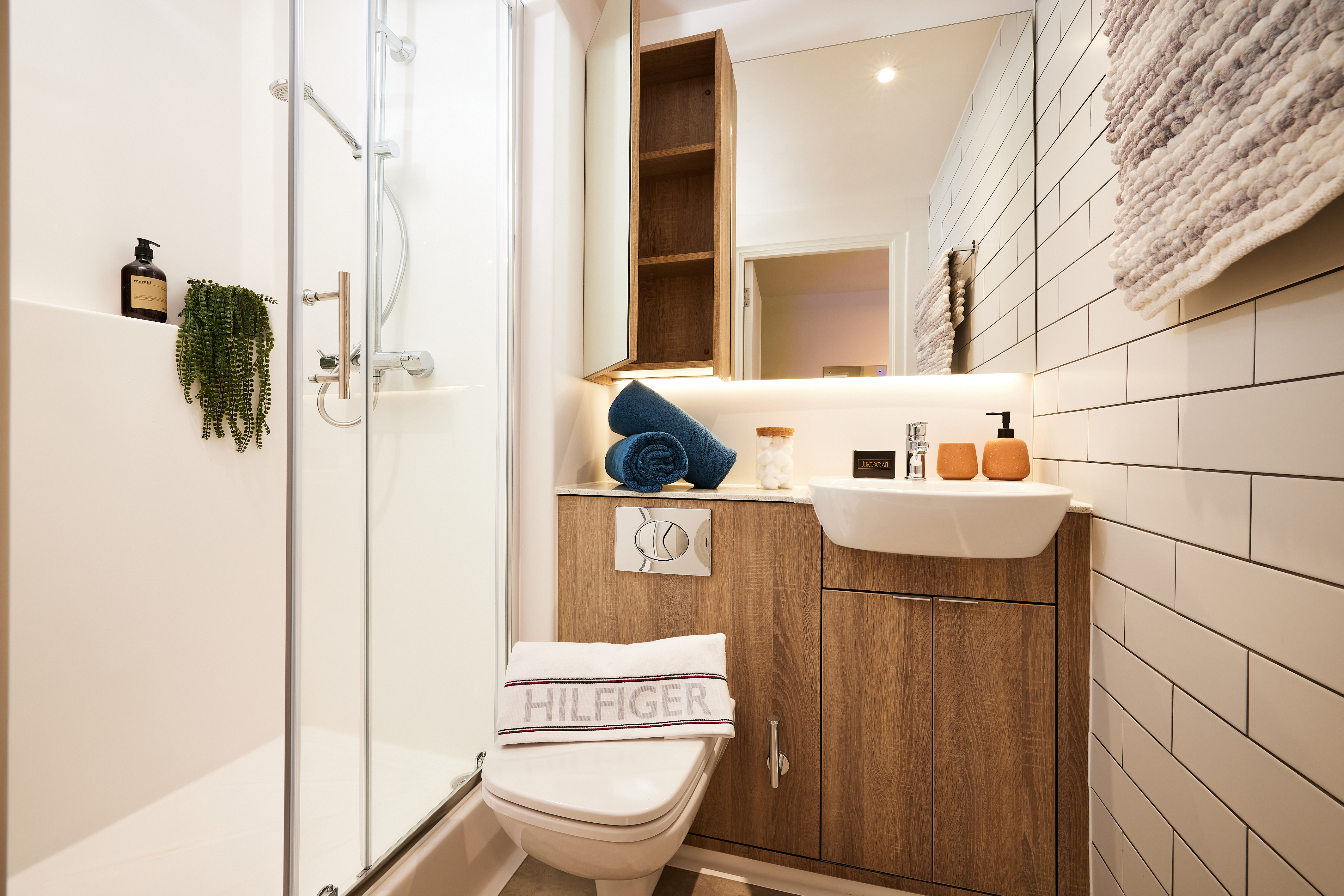 A modern bathroom with a glass shower, wooden cabinetry, and a white sink. A blue towel and toiletries are on the counter. A potted plant hangs in the shower. A towel with "HILFIGER" sits on the toilet lid, reminiscent of Cannon Park's sleek style.
