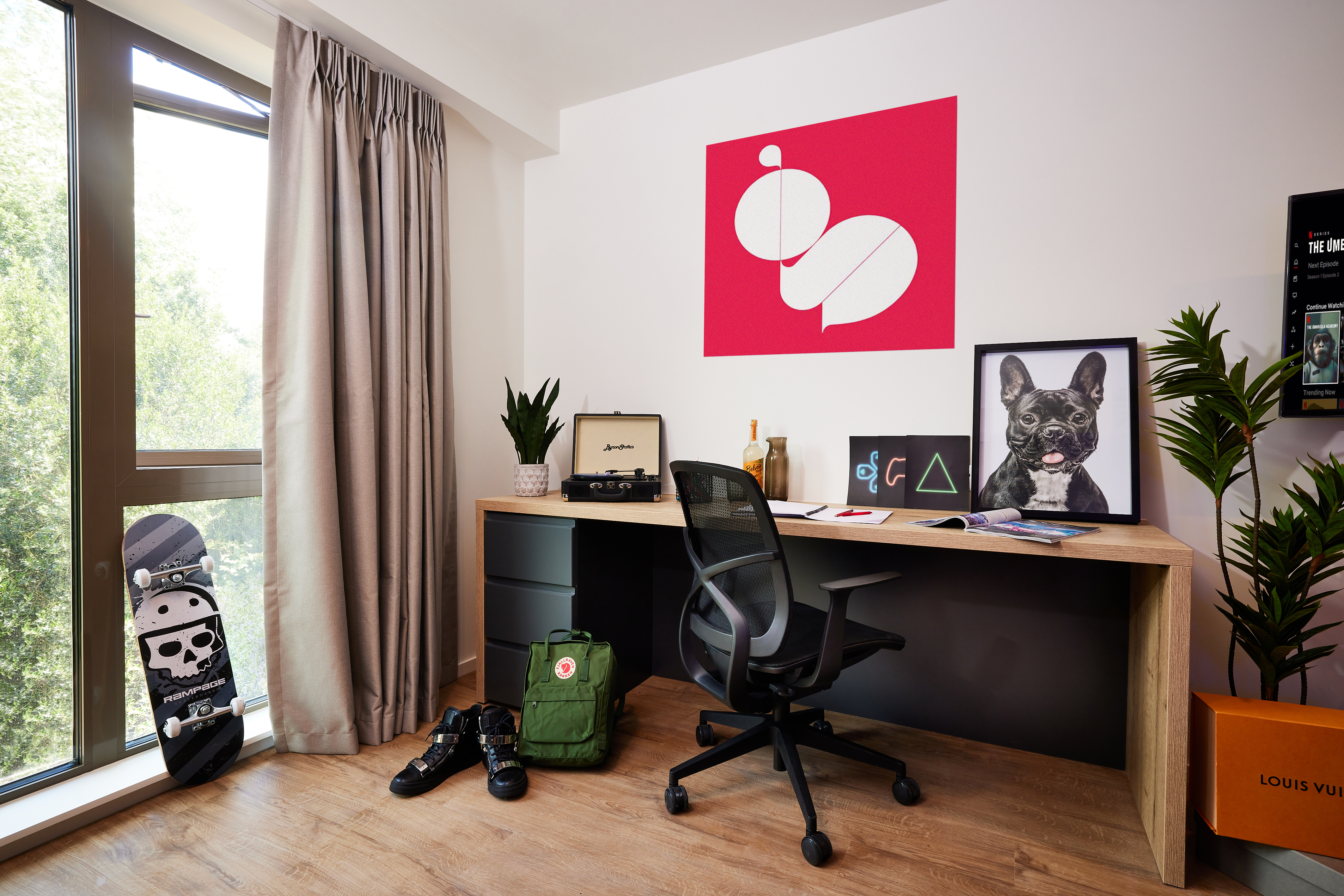 A modern bedroom in Warwick features a wooden desk with a computer, books, and wall art. A black chair and green backpack are nearby. A skateboard rests by the window, draped with beige curtains. The walls boast a pink abstract painting and pet artwork reminiscent of Cannon Park's vibrant spirit.