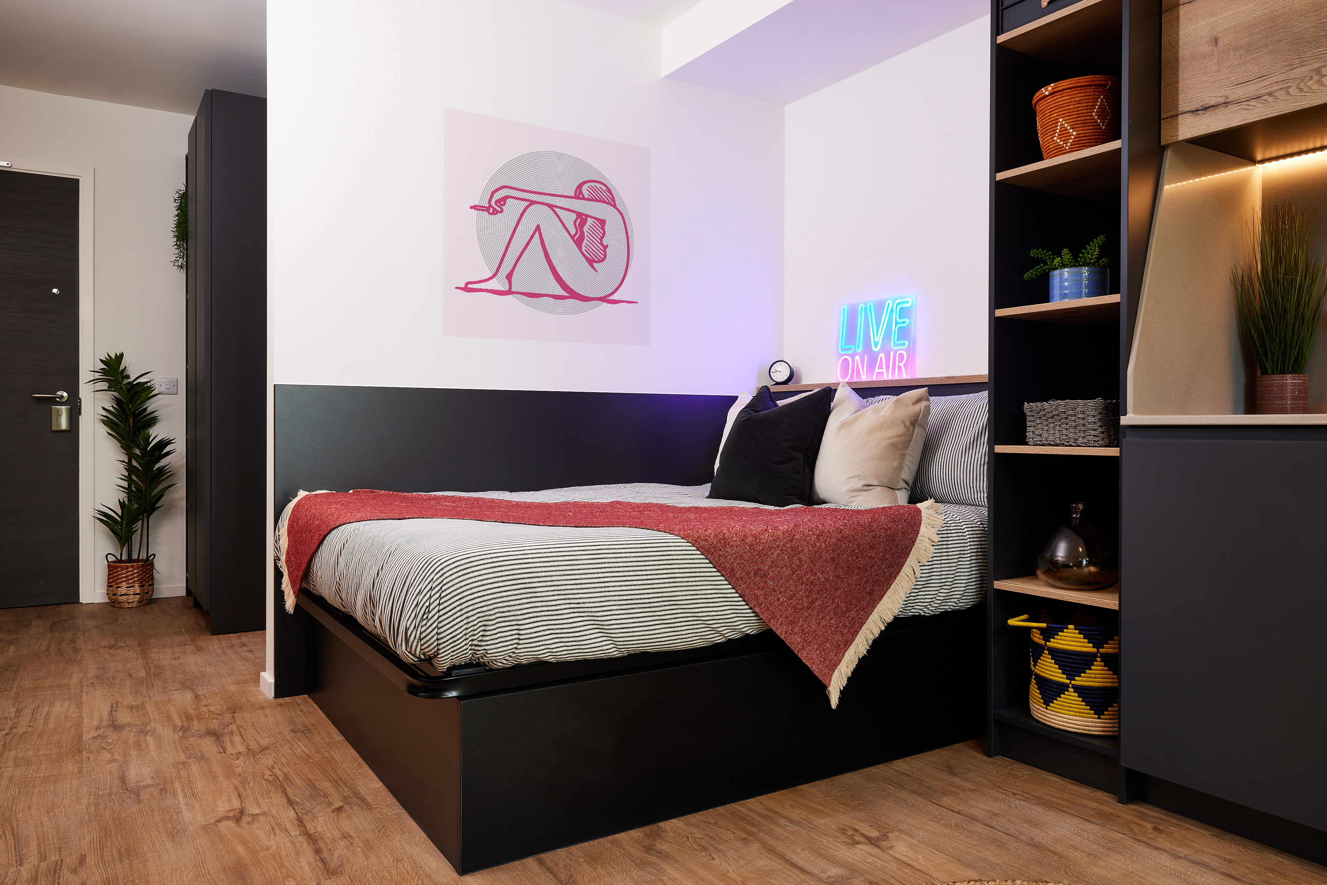 Modern bedroom with a neatly made bed featuring a red throw blanket and striped bedding. A neon "LIVE ON AIR" sign glows on a shelf beside the bed, bringing lively energy reminiscent of Warwick's vibrant Cannon Park. Potted plants decorate the room, and art hangs on the wall above the bed.
