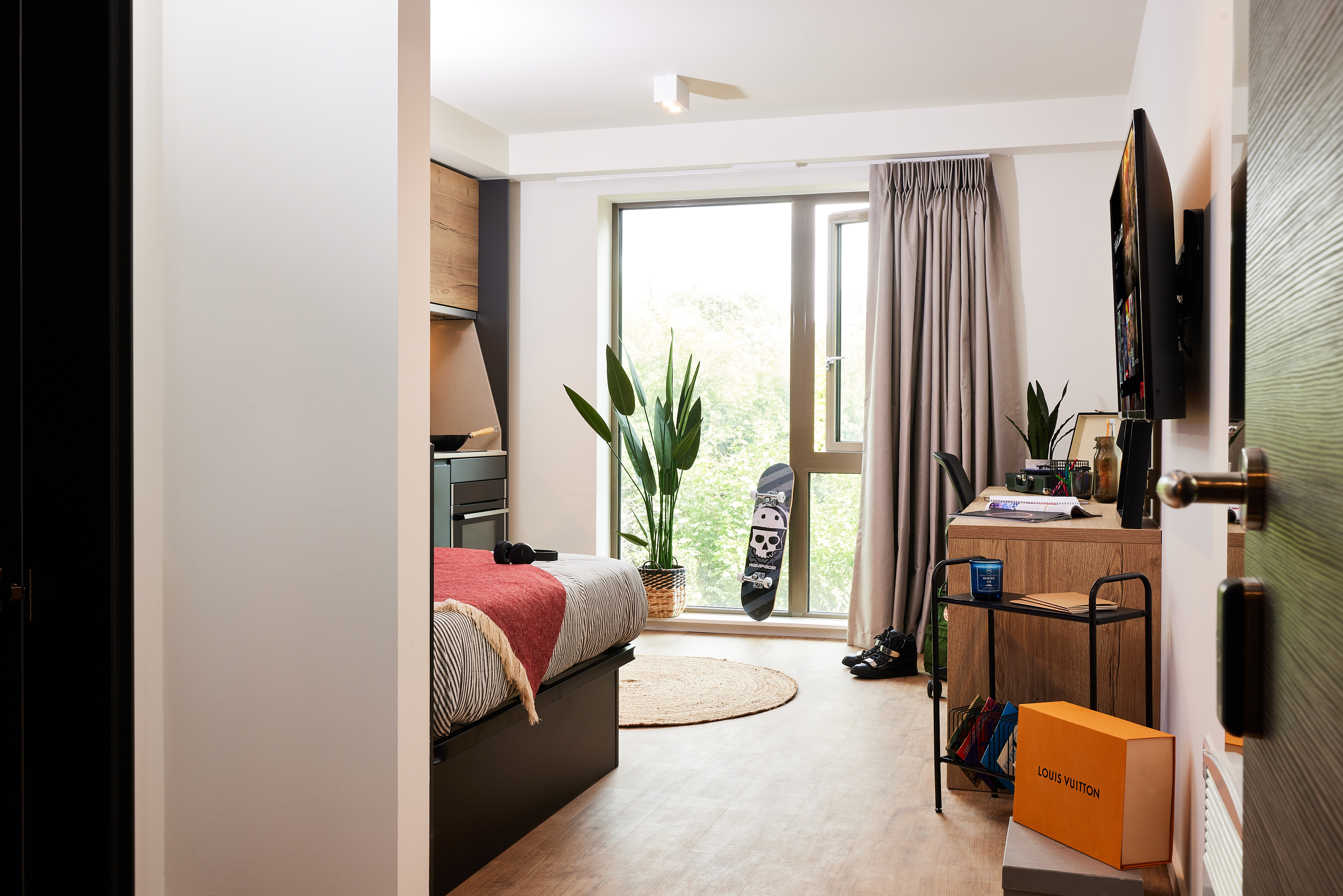 A modern, well-lit bedroom with a large window overlooking Warwick. The room features a bed with a red throw, plants, a skateboard with a skull design, and a wall-mounted TV. A shelf holds books, while on the floor rests an iconic Louis Vuitton box reminiscent of Cannon Park's chic style.