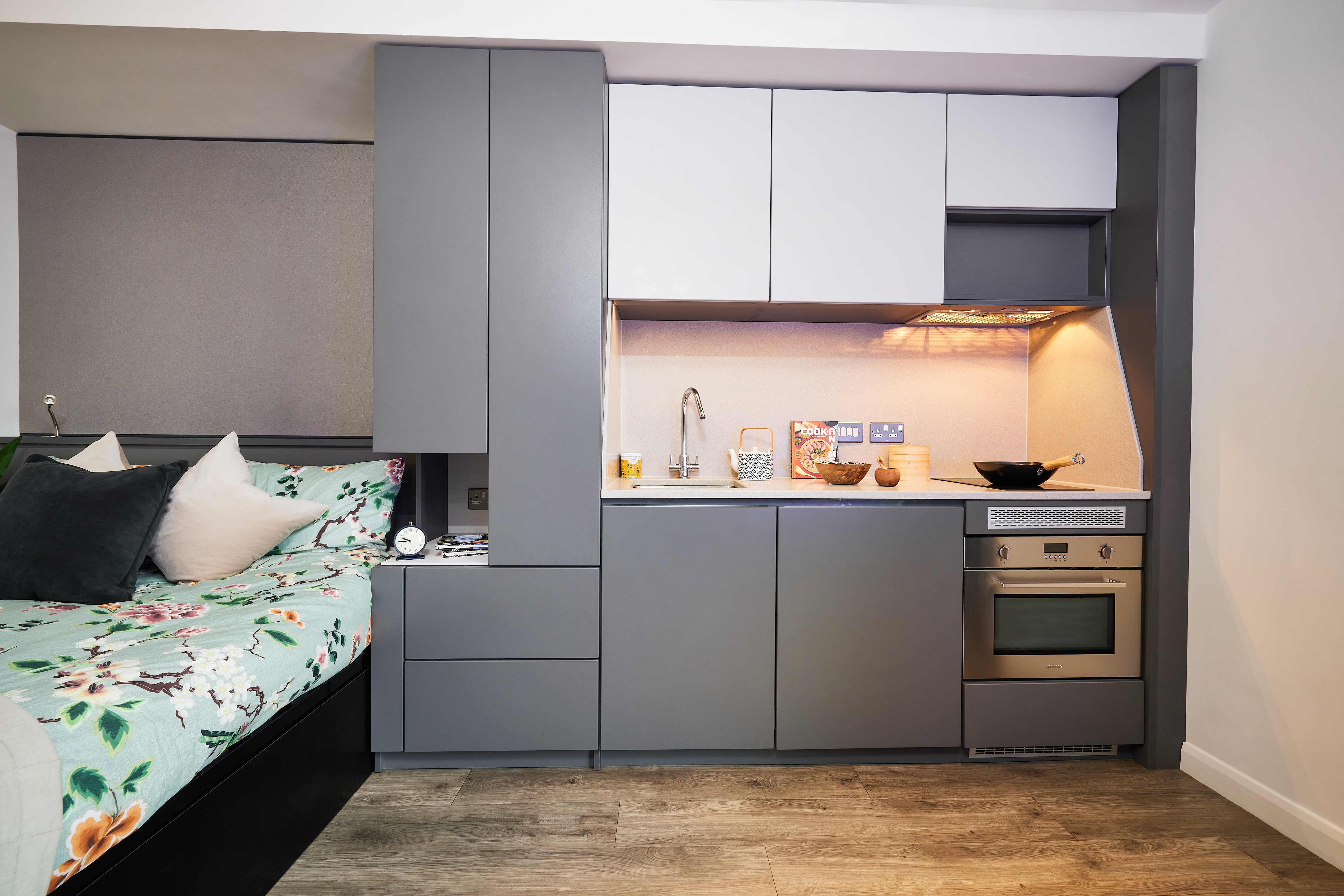 Discover a compact modern studio apartment on Strawberry Place with a kitchenette featuring gray cabinets, a small stovetop, and oven. The bed with floral bedding lies adjacent to the kitchen area. Wooden floors and minimal decor make for a cozy, efficient living space.