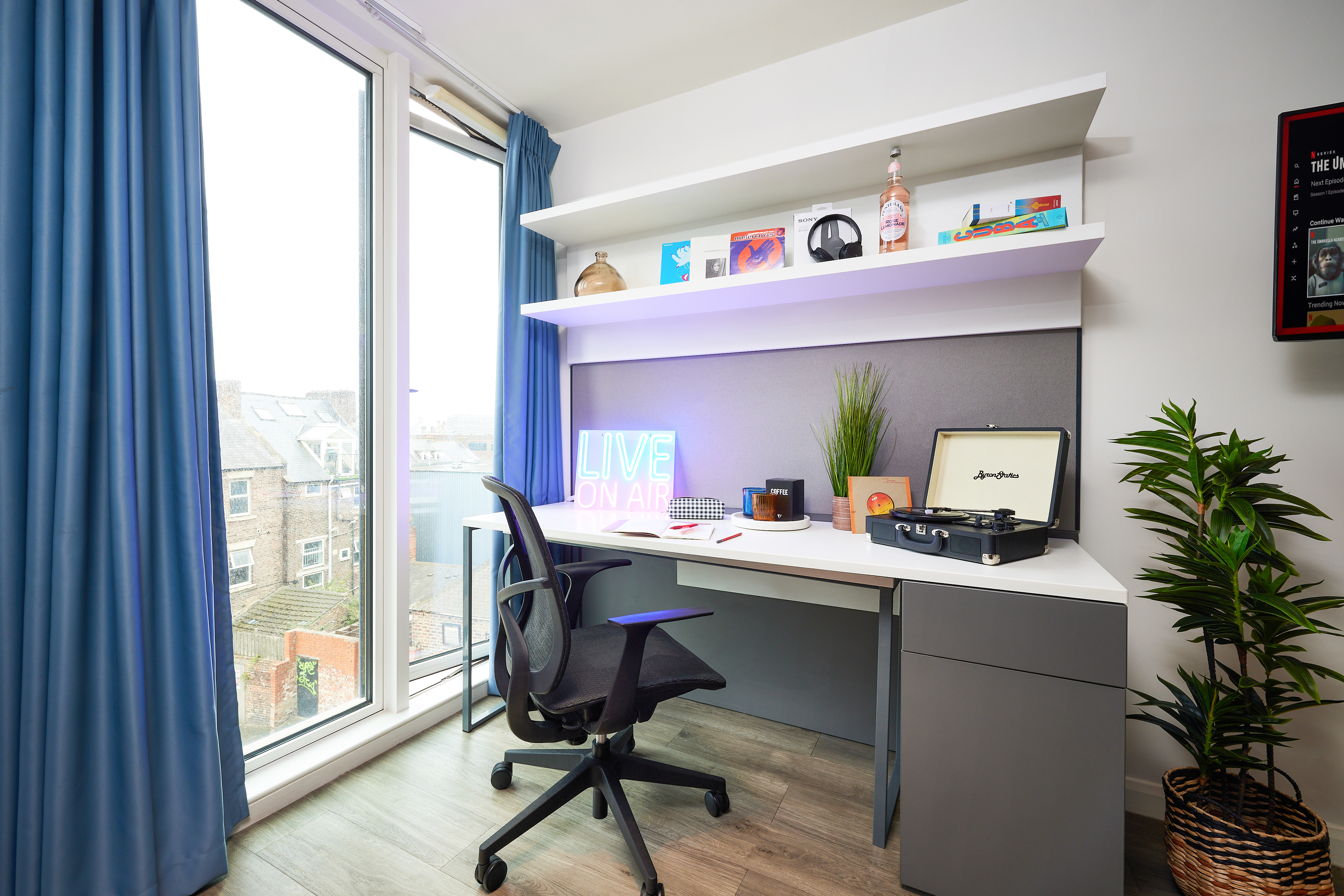 A modern home office features a sleek desk, ergonomic chair, and floating shelves. A neon "LIVE ON AIR" sign radiates on the desk next to a vintage record player. Large windows with blue curtains frame rooftop views, while lush plants add greenery to this serene place.