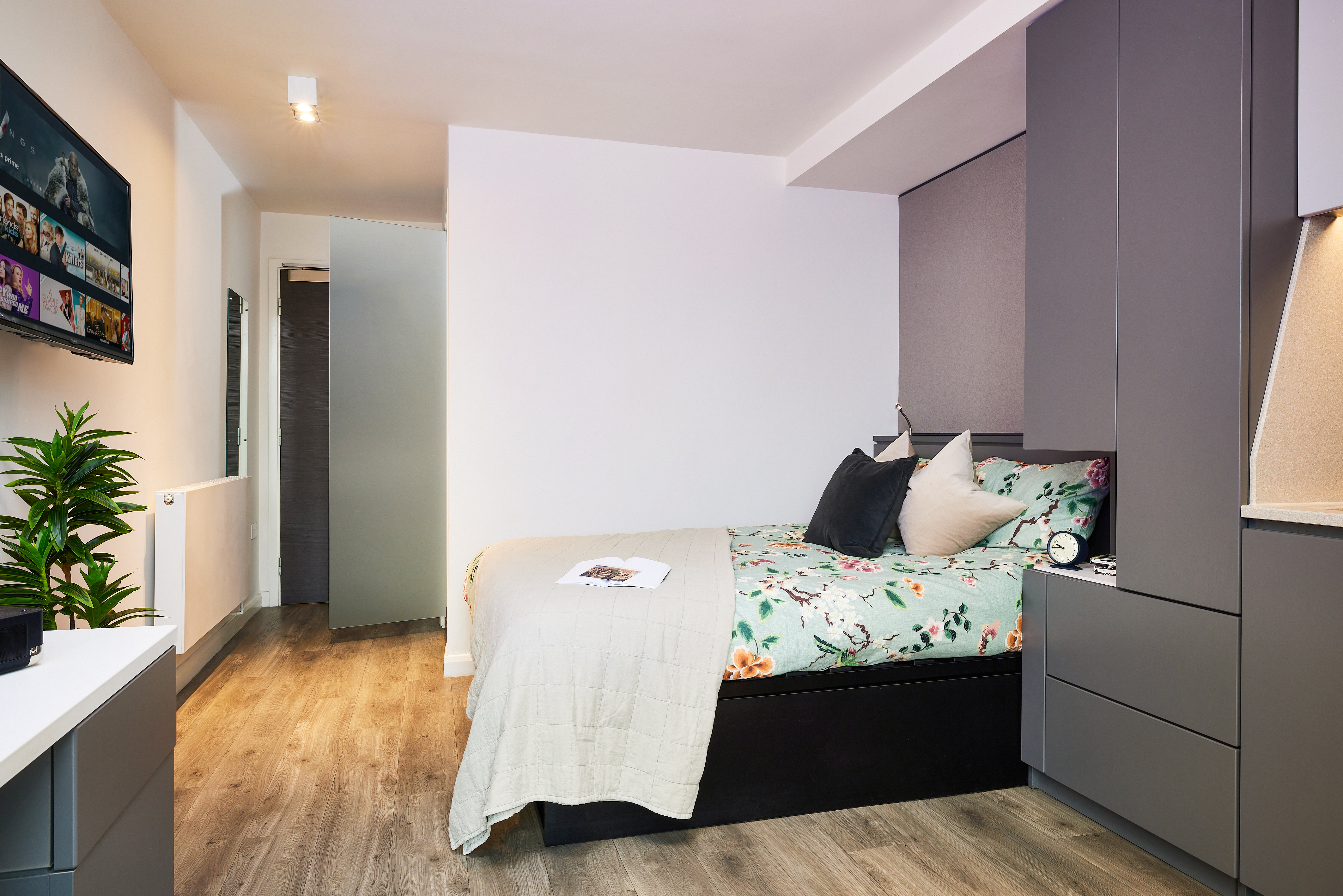 In the modern bedroom at Strawberry Place, a neatly made bed with floral bedding is surrounded by sleek gray furniture. A small green plant sits on a white desk, while a flat-screen TV is mounted on the wall. The wooden floor enhances the room's warm ambiance.