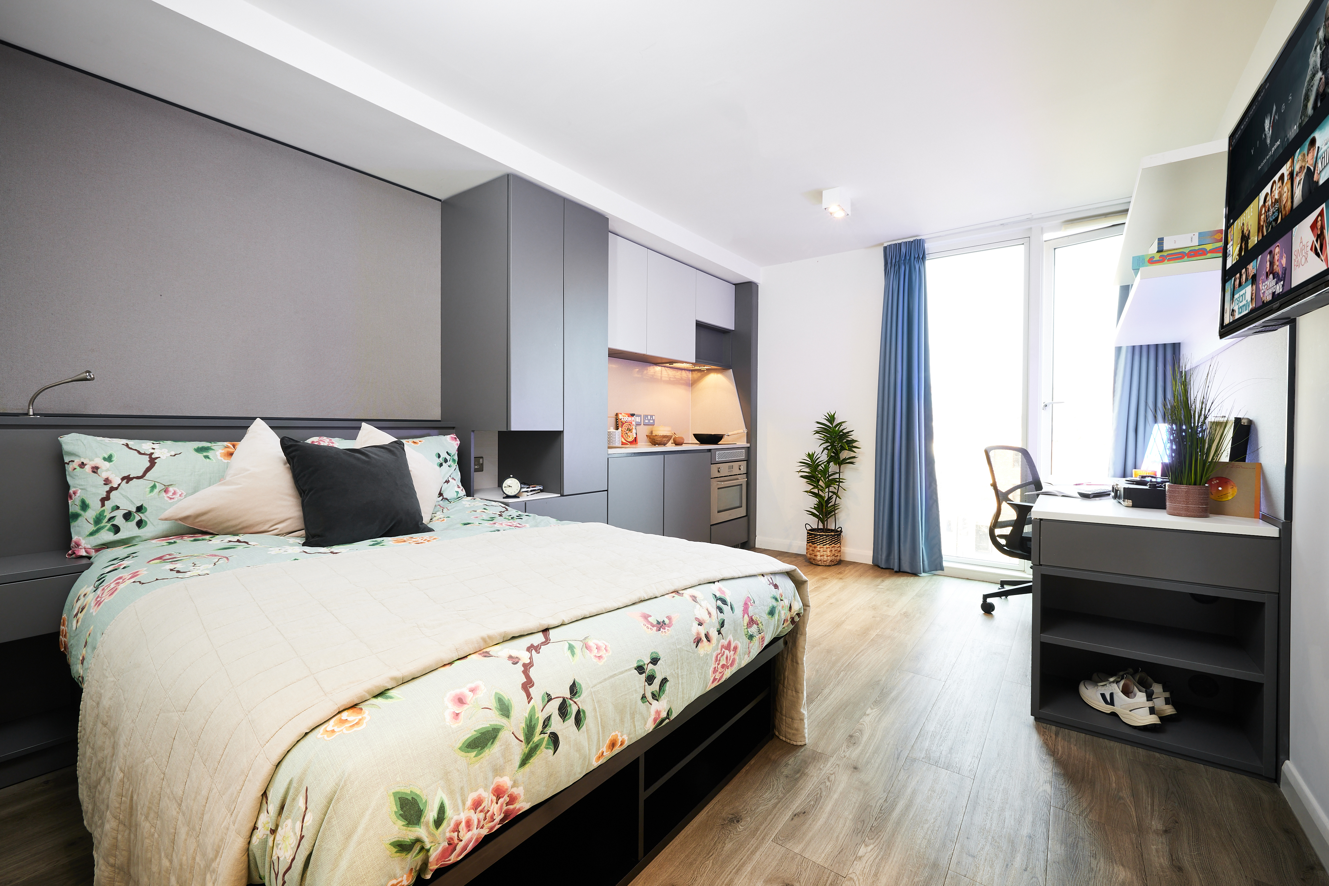 A modern bedroom at Strawberry Place features a floral-patterned bed, built-in grey cabinets, and a kitchenette area. A desk with a computer and books sits by the window adorned with blue curtains. A small plant adds greenery to the space.