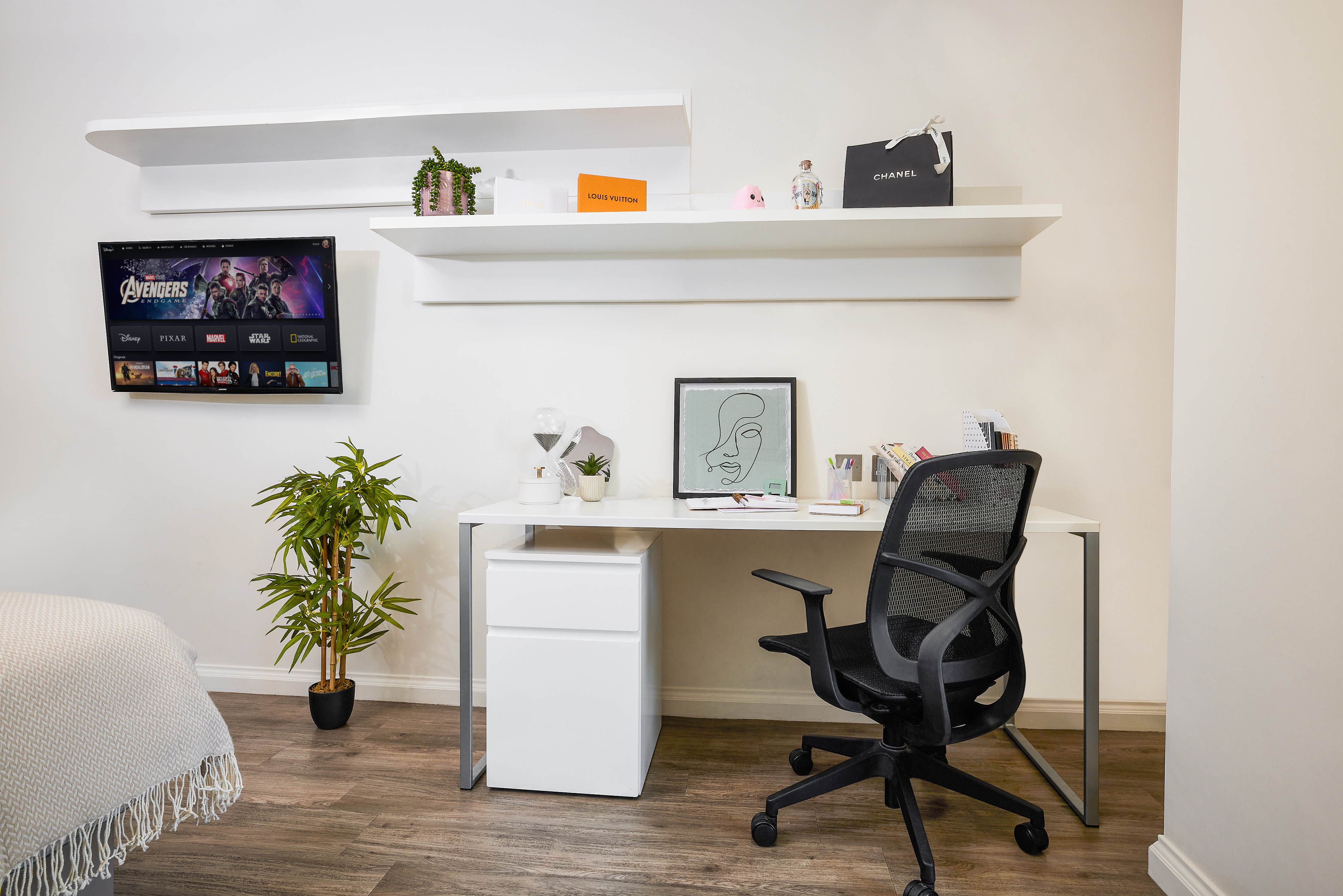 Minimalist home office with a white desk, black chair, and potted plant. Wall-mounted TV displays a streaming platform. Shelves hold plants and decor. Desk features a framed line drawing, small fan, notebooks, and stationery. Light wooden floor.