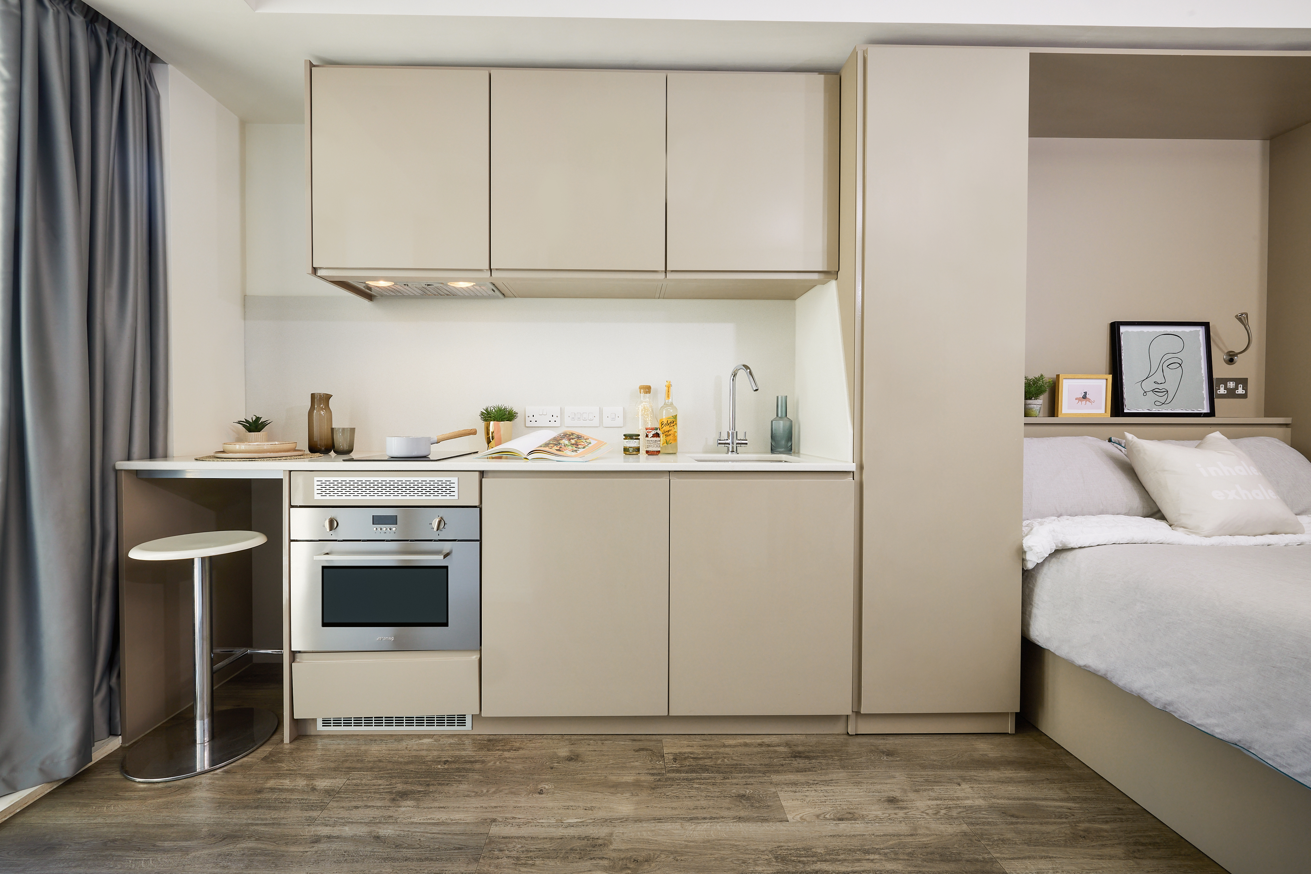 A compact studio apartment with a small kitchen featuring beige cabinets, a stove, and a countertop with a stool. Next to the kitchen is a bed with white bedding. A small plant and framed art pieces are placed throughout the room.