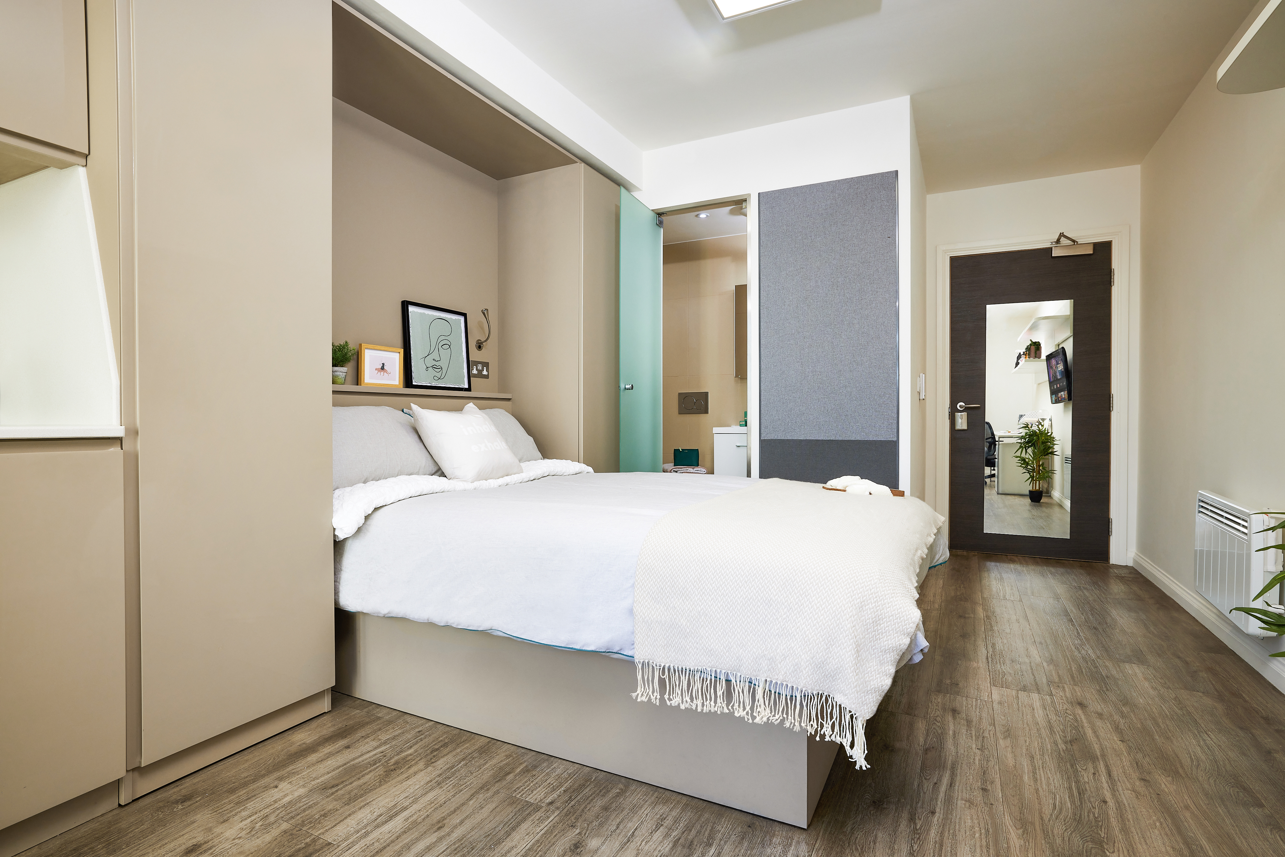 A modern bedroom with a neatly made bed adorned with white pillows and a beige throw. The room features a wooden floor, minimalistic decor, a small picture on the wall, and an open bathroom door with visible fixtures.