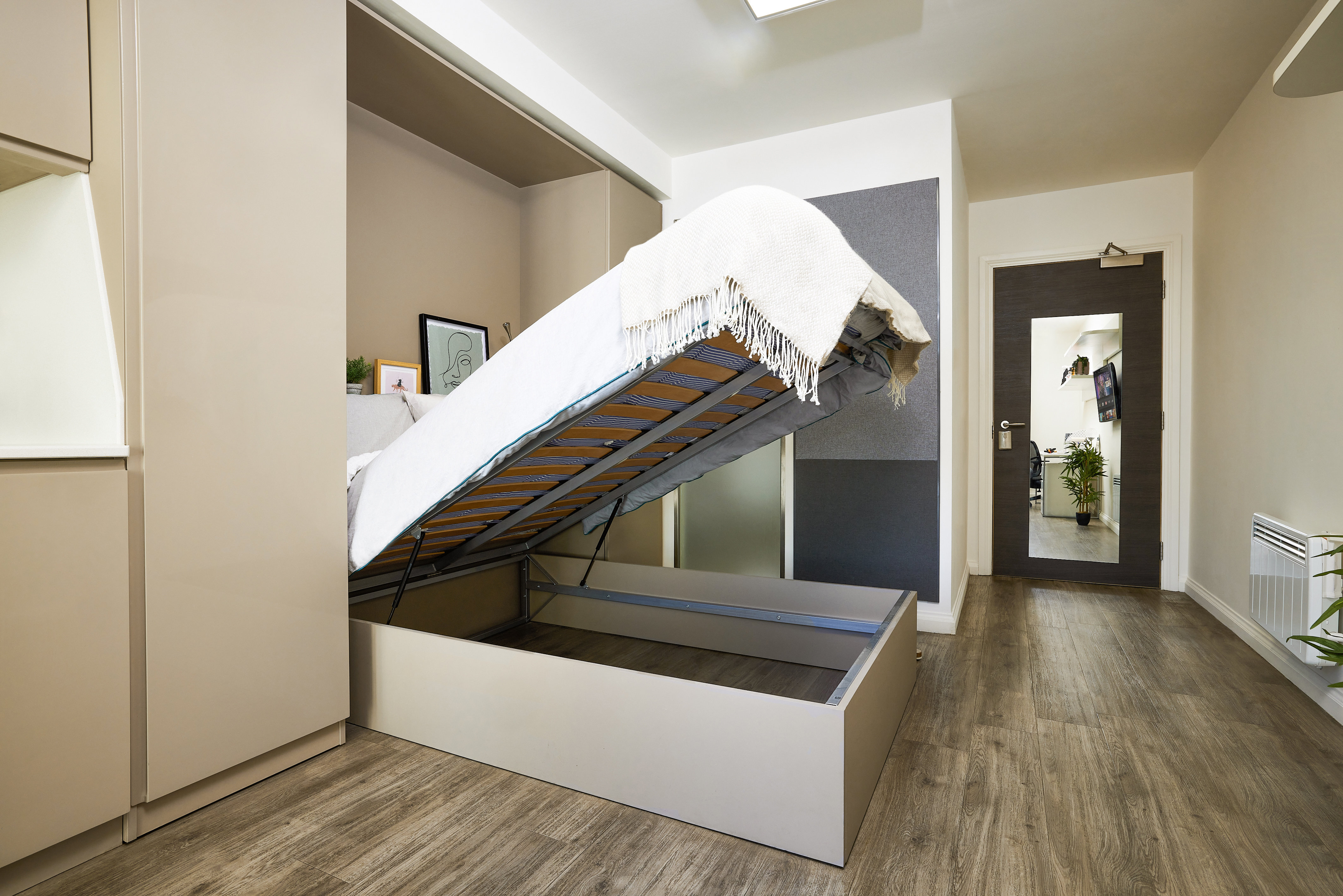 A modern bedroom with a raised bed revealing storage space underneath. The room features wooden flooring, neutral-colored walls, and minimalistic decor, including a plant and artwork. A door and a hallway mirror are visible in the background.