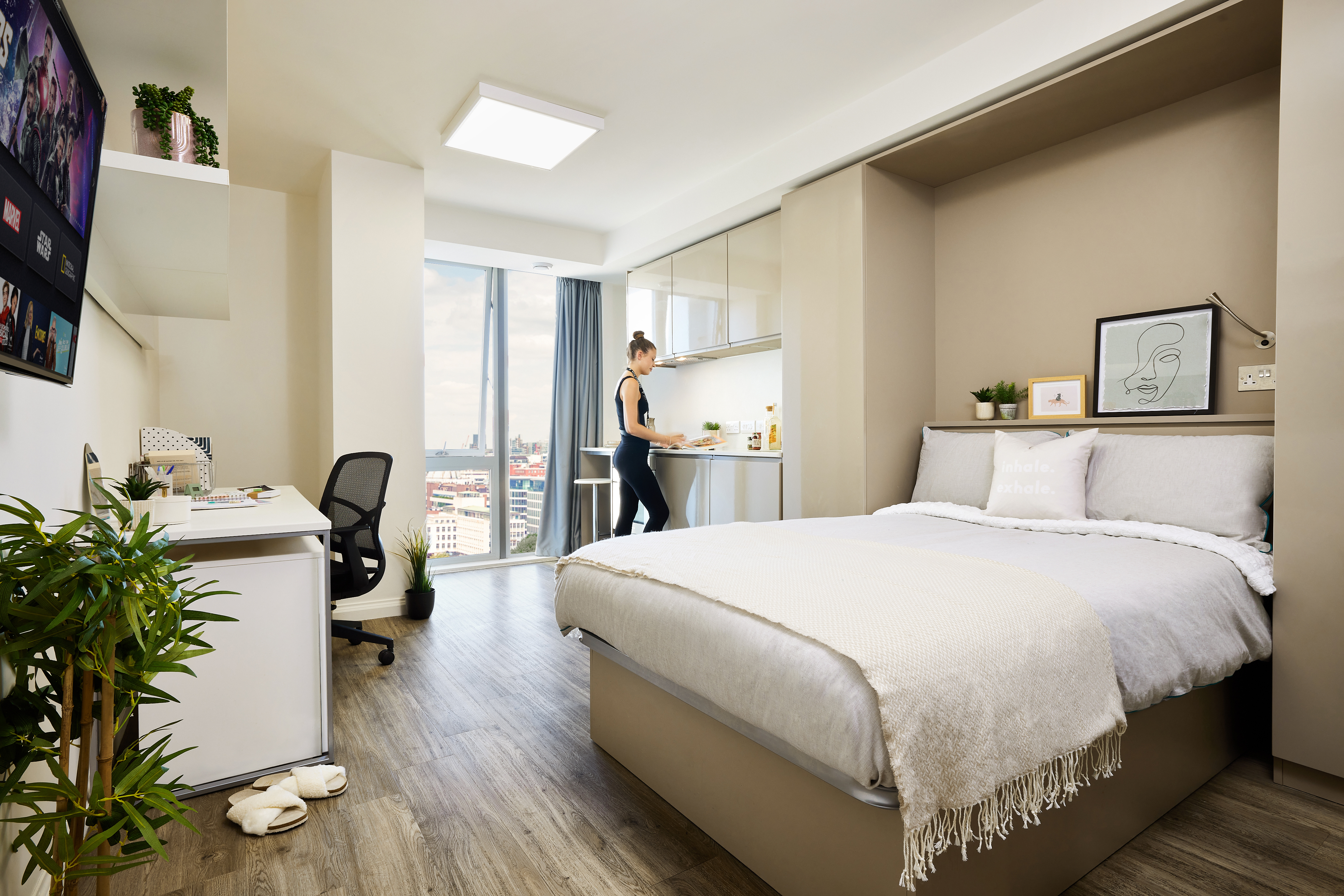 Modern studio apartment with wood flooring. Includes a bed with white linens, framed art above, a desk with chair, TV, and plants. A person is cooking in a kitchenette with large windows showing a city view. Bright and spacious interior.