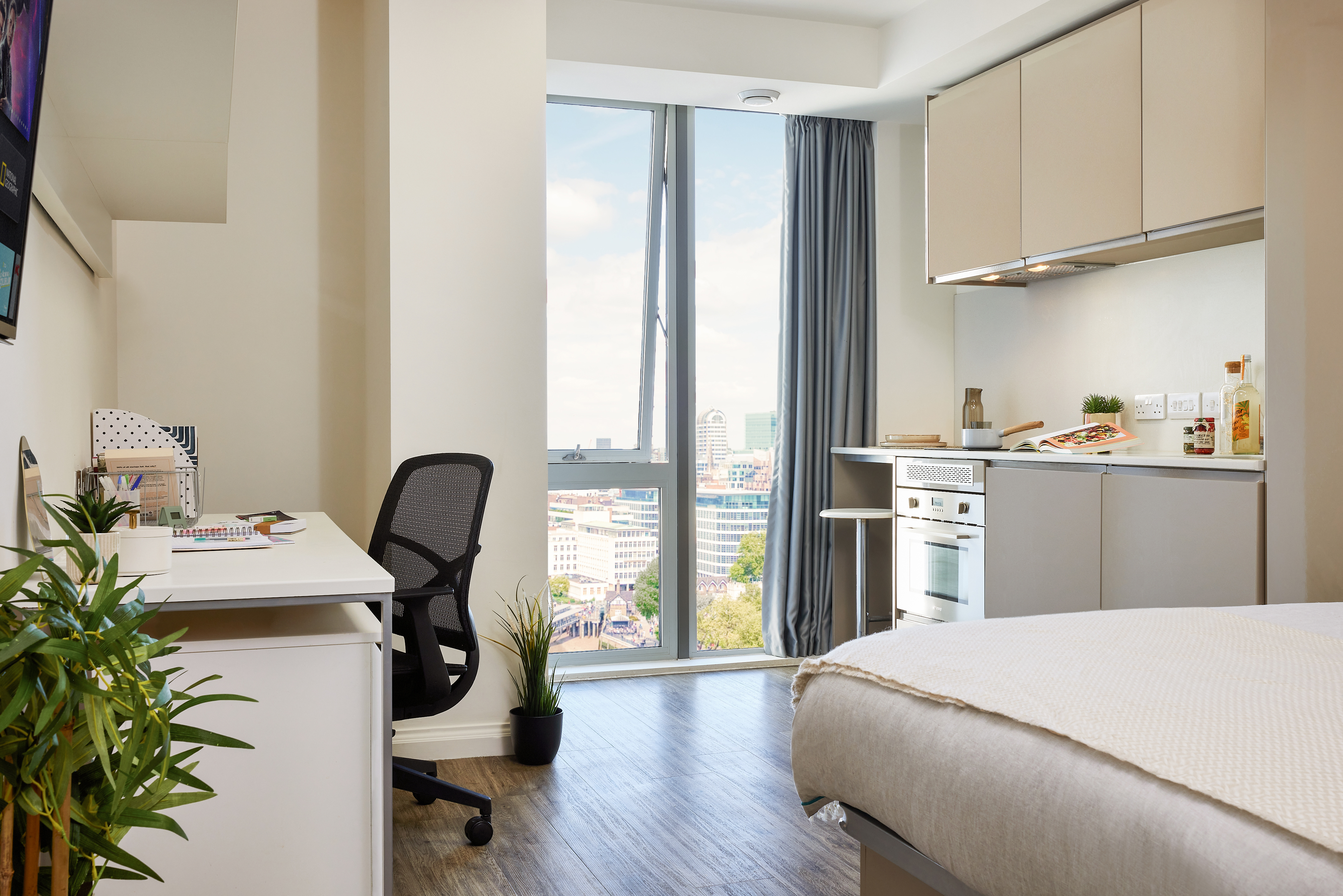 Modern studio apartment with a bed, desk, and office chair. There's a kitchenette with a stove, cabinets, and a small plant on the counter. A large window offers a city view. Light wood flooring and a cozy, organized layout.