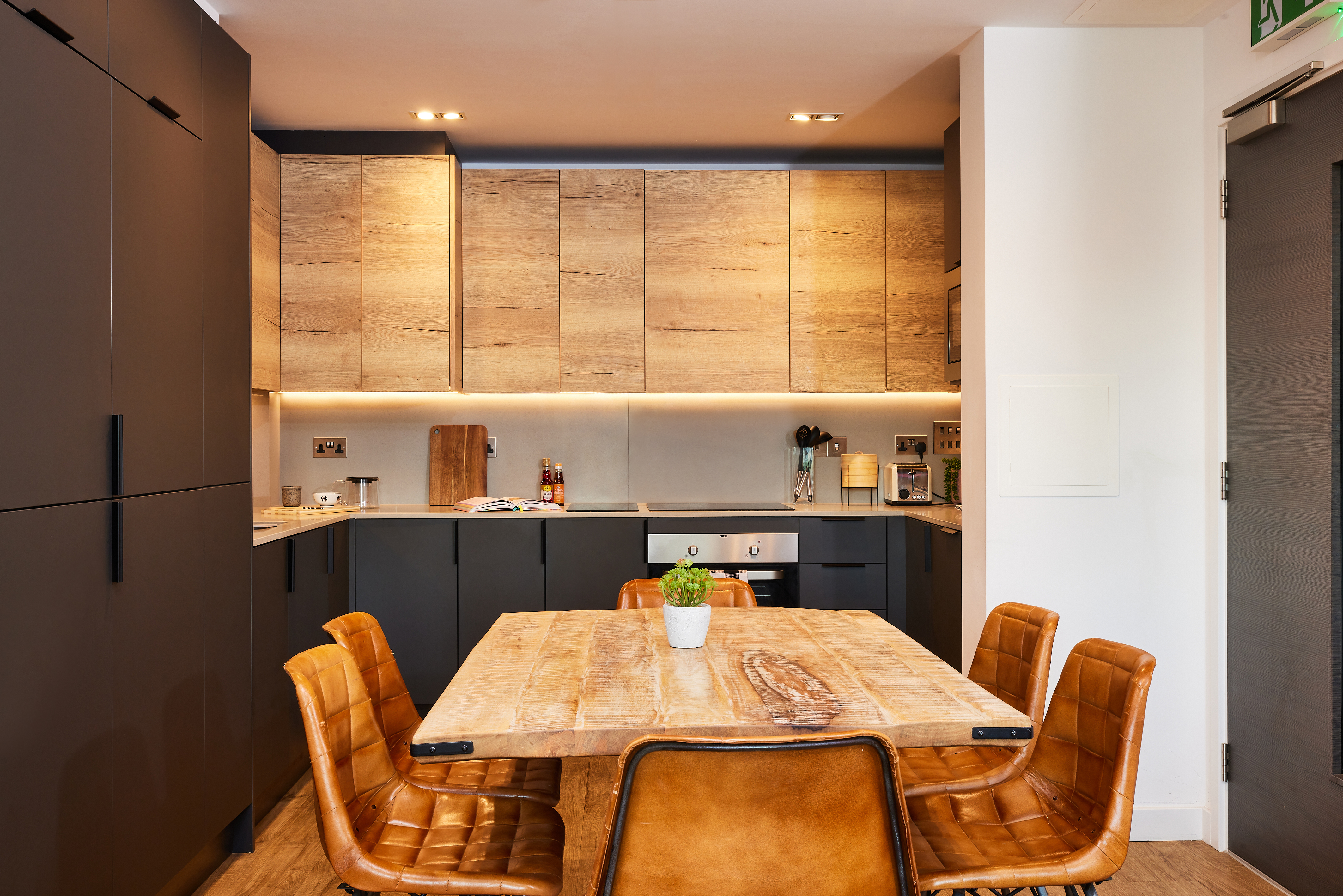 Cozy kitchen with wooden cabinets and a central dining table surrounded by four brown leather chairs, perfect for your Lewisham real estate dreams. The counter has kitchenware, and a small plant graces the table. Warm lighting enhances the inviting atmosphere.