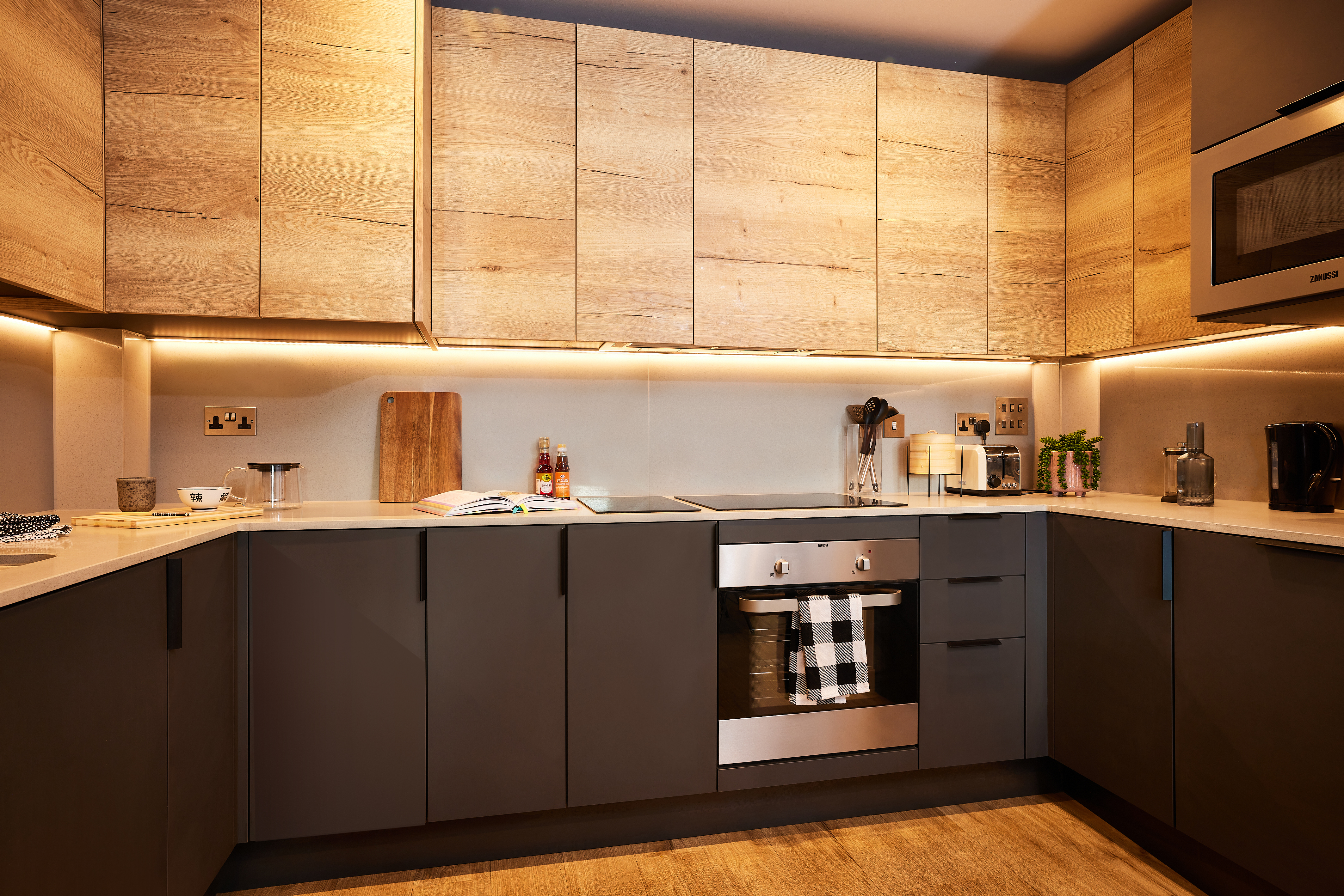 Located in the Lewisham Exchange, this modern kitchen features wood and dark cabinets, stainless steel appliances, and under-cabinet lighting. A tea towel hangs on the oven door, accompanied by a cutting board and herbs on the countertop. Wood flooring completes the look.