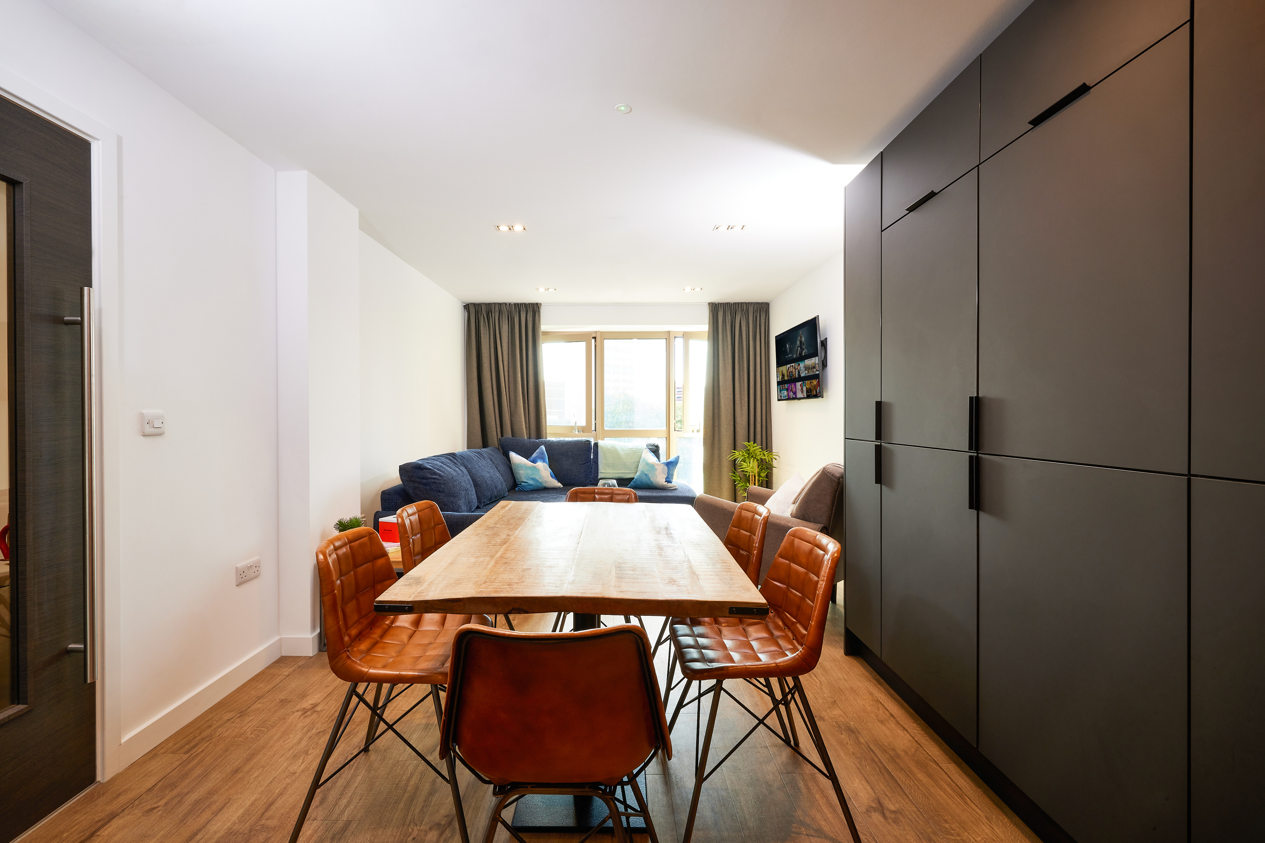 A modern living and dining area at Lewisham Exchange features a wooden table surrounded by brown leather chairs. A blue sofa against the wall with cushions sits next to large windows with curtains. A sleek, dark storage unit lines one wall, while a TV hangs nearby.