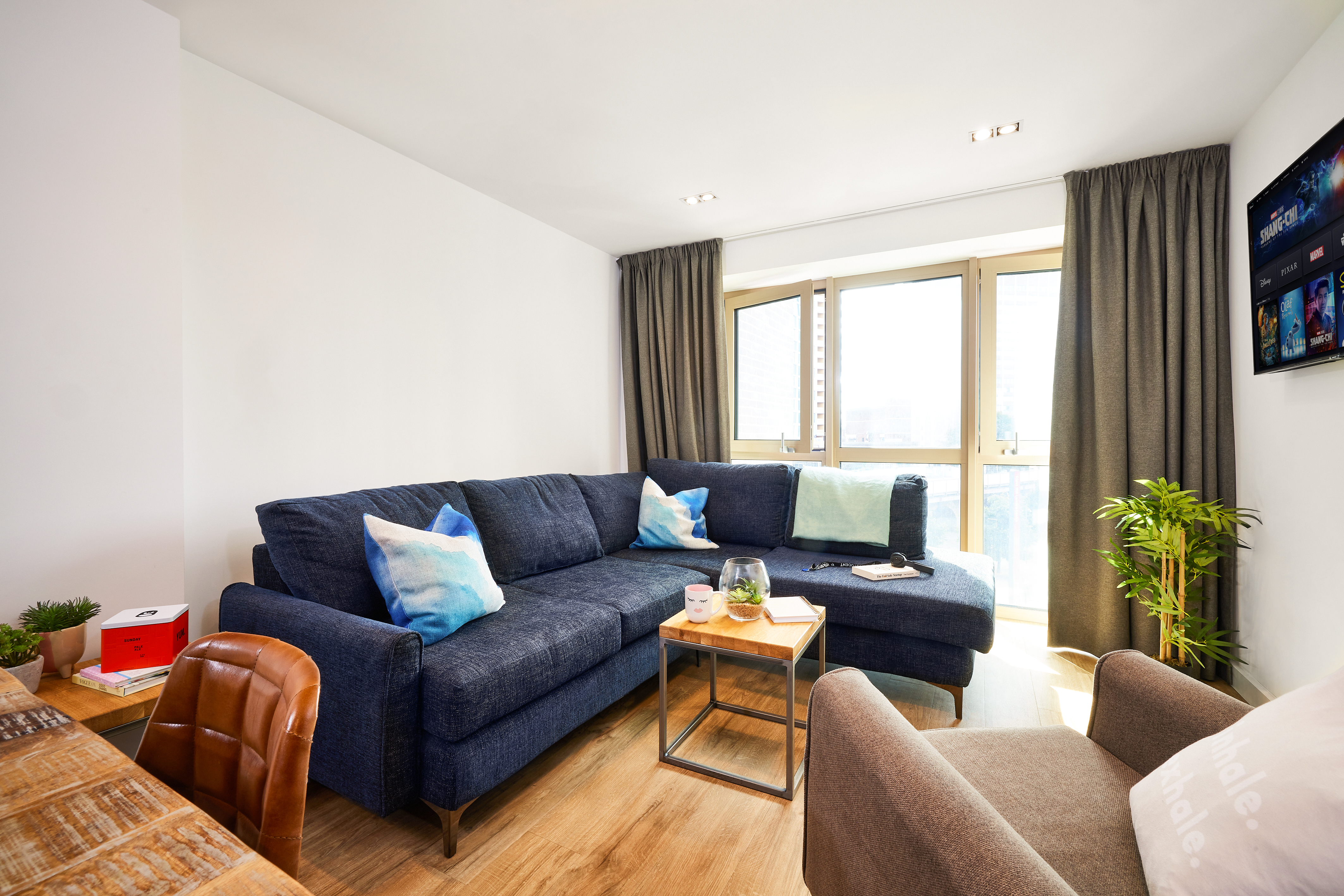 In the heart of the Lewisham Exchange, this modern living room features a dark blue sectional sofa, light wood flooring, and a small wooden coffee table. A potted plant sits in the corner near large windows with gray curtains, perfectly complementing the wall-mounted TV's colorful display.