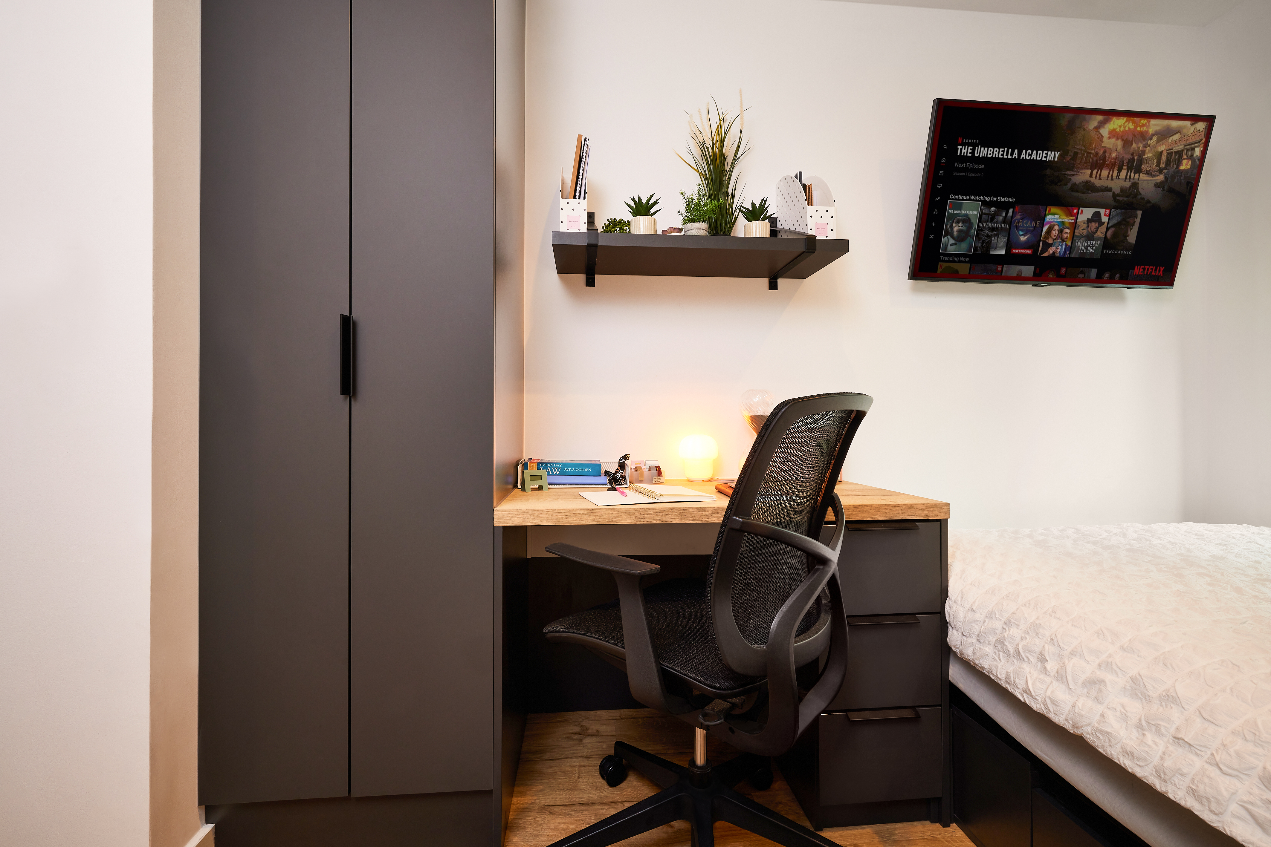 A modern bedroom in Lewisham boasts a black wardrobe, a wooden desk with an office chair, and a shelf adorned with plants and stationery. A TV on the wall displays a streaming app, while the bed is dressed with a white textured comforter.