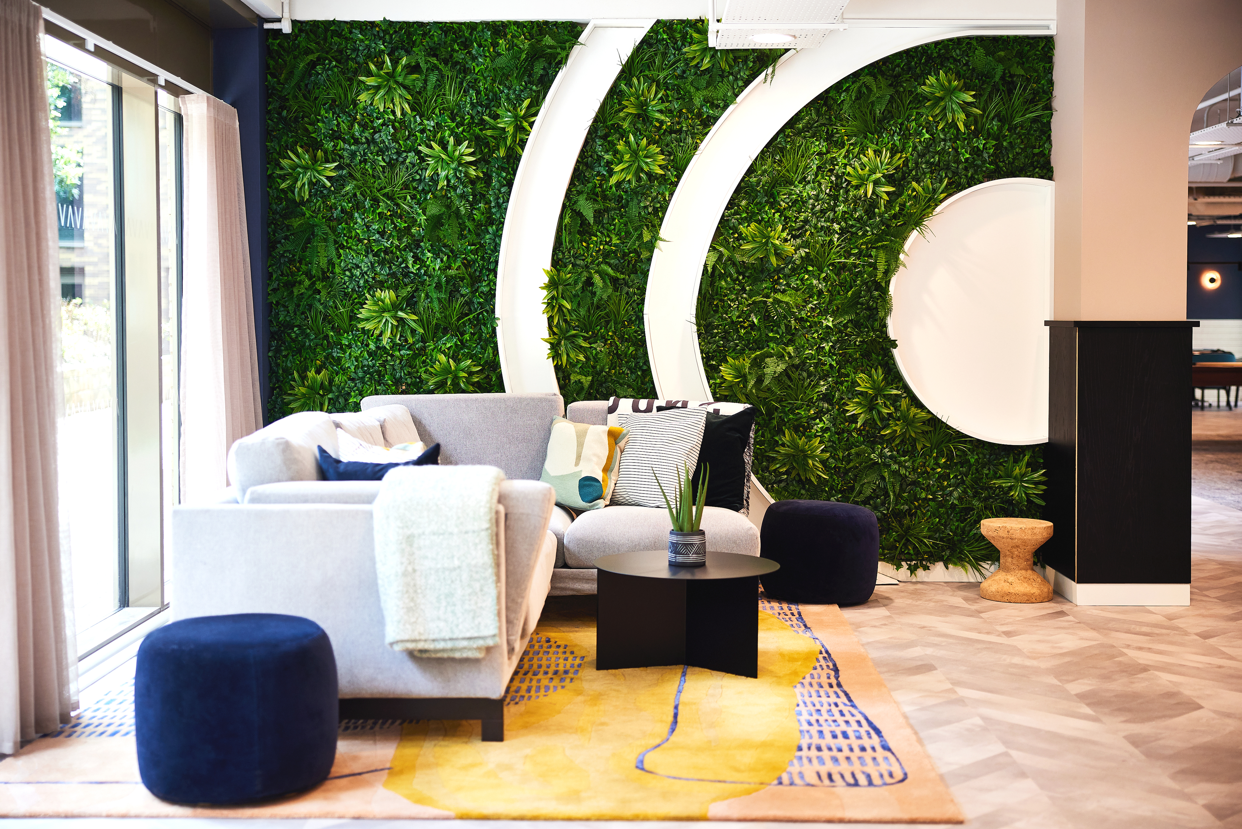 In the modern lounge area near Cannon Park, a gray sofa complements navy ottomans and a black table on a yellow geometric rug. The backdrop features a lush green vertical garden with circular white accents, while a large window on the left invites natural light into this Warwick space.