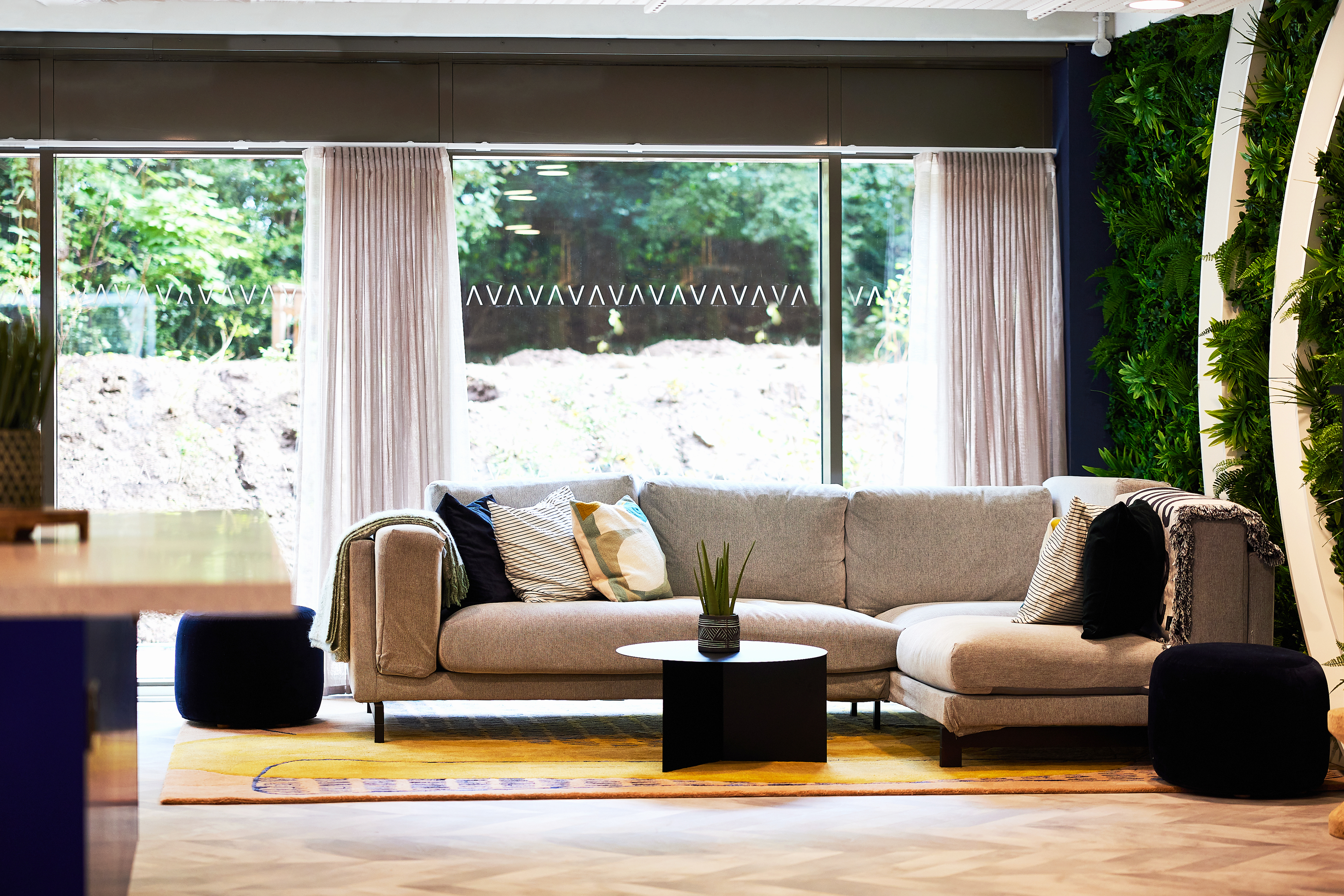 A modern living room at Warwick features a light gray sectional sofa with pillows. A wooden round coffee table sits in front, with a plant on it. Large windows behind reveal a lush outdoor view, and vertical green plant walls flank the scene, echoing the tranquility of Cannon Park.