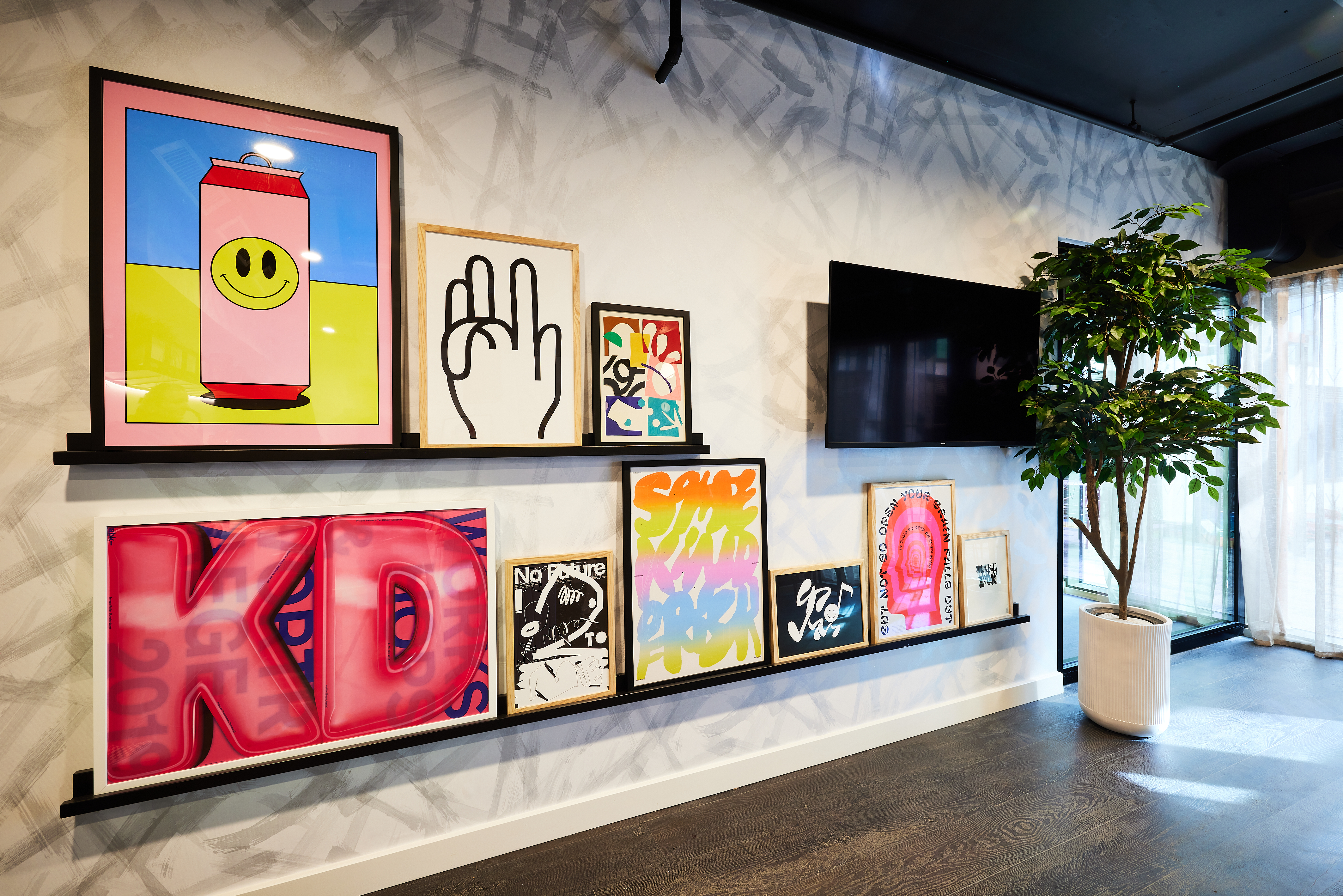 A modern interior in a Cannon Park home features colorful pop art paintings, including a pink can with a smiley face, a hand gesture, and bold lettering. A flat-screen TV and potted plant add charm to the space with dark flooring and a geometric ceiling pattern.
