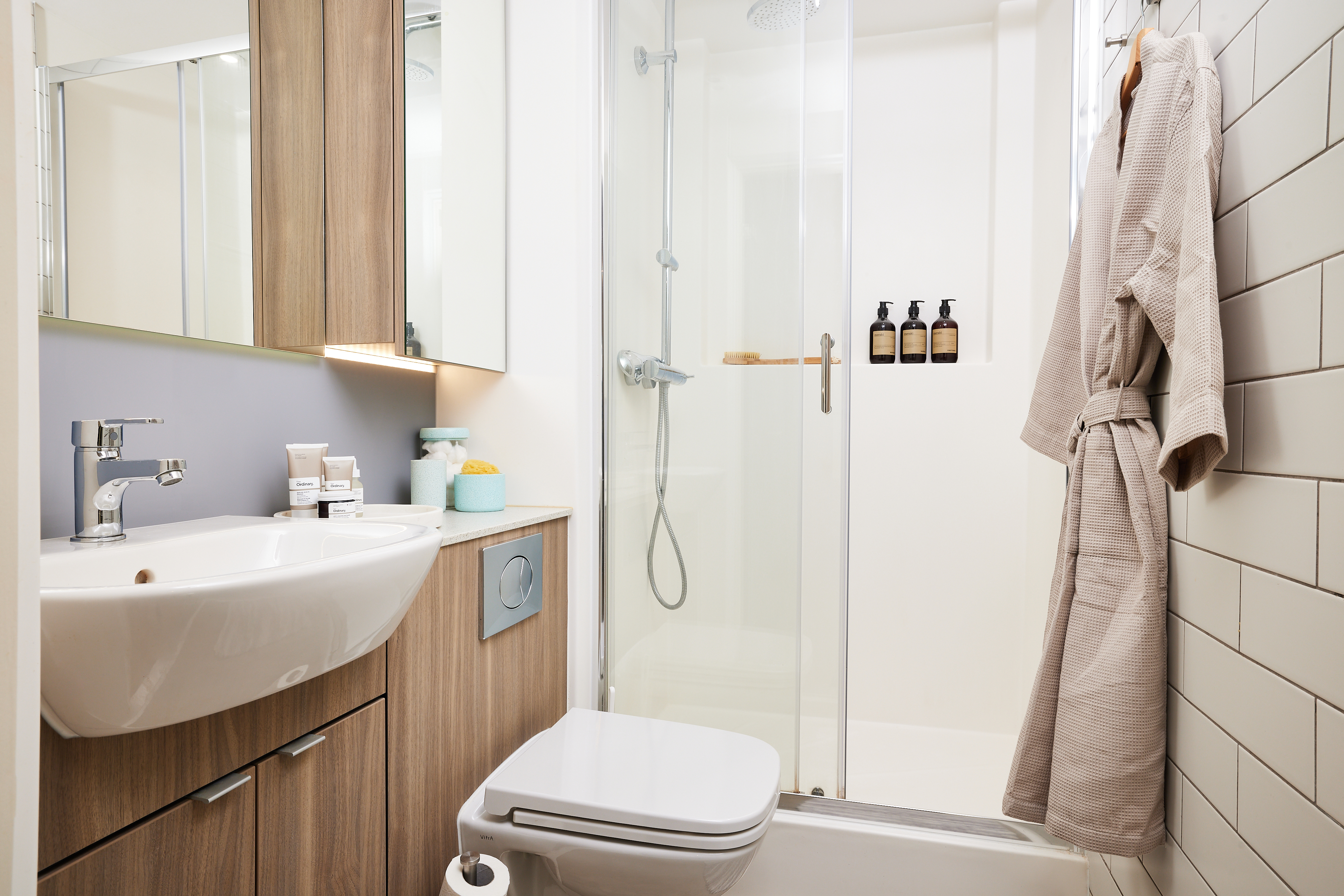 Modern bathroom with a glass-enclosed shower, wooden cabinets, a white sink, and a wall-mounted toilet. A beige robe hangs on the tiled wall, and toiletries are neatly arranged on the counter and a shelf in the shower.