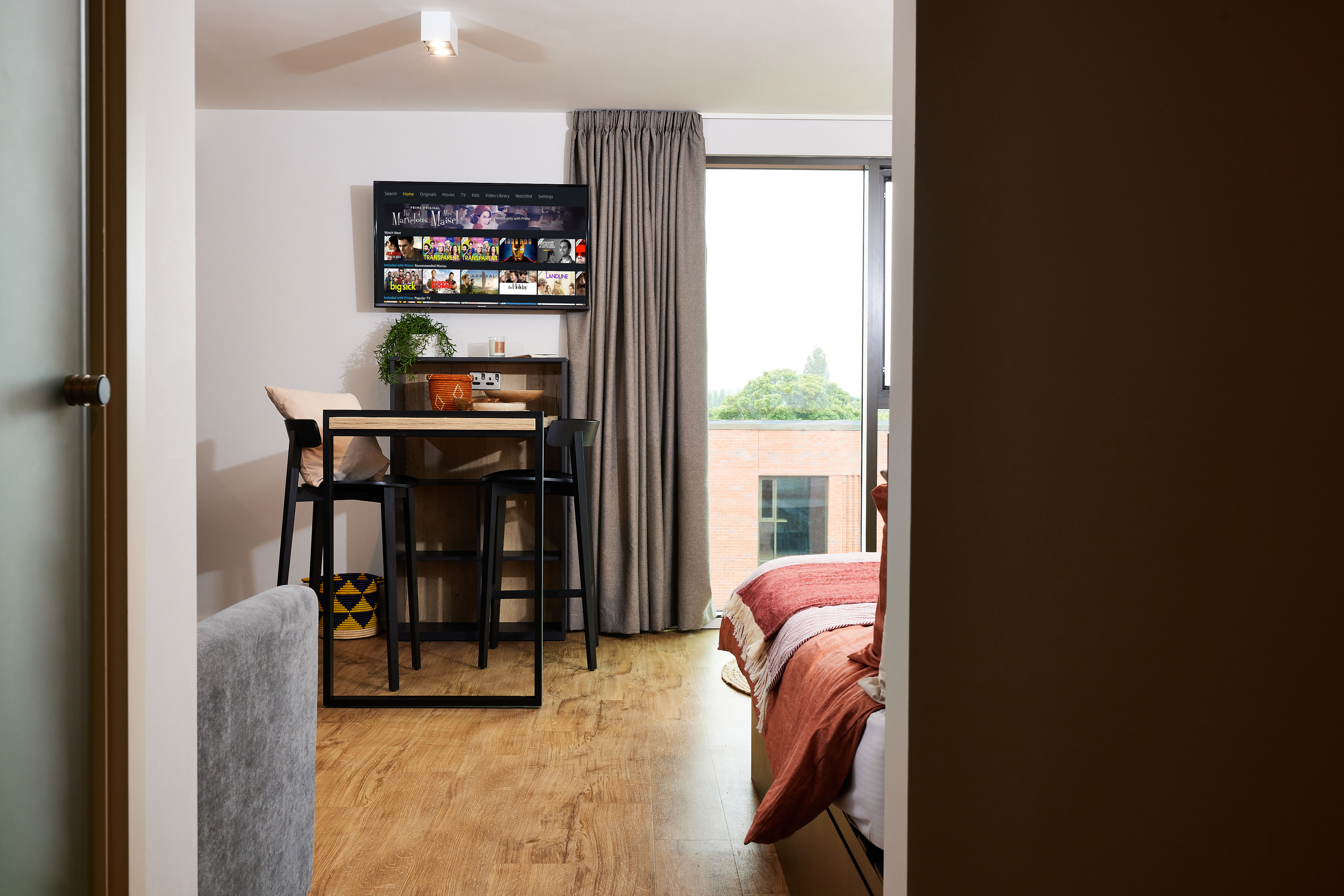 A modern studio apartment featuring a cozy bed with orange bedding, a small dining table with two chairs, a wall-mounted TV, and a large window with curtains. The room has wooden flooring and a view of some trees outside.