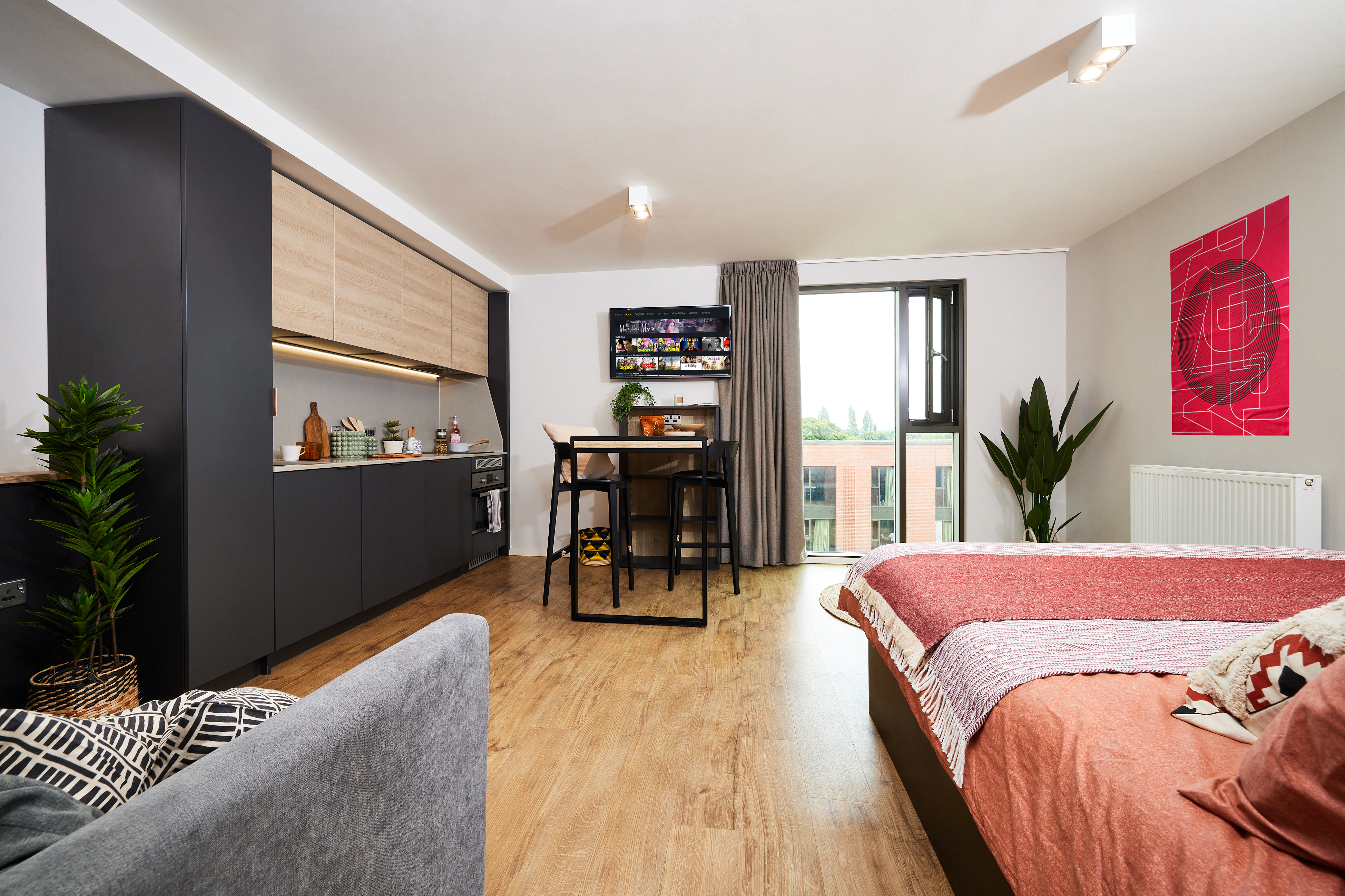 Modern studio apartment with a small kitchen, a table with chairs, and a bed. There are potted plants, a large window, wooden flooring, and wall art. The decor features neutral colors with splashes of red accents.