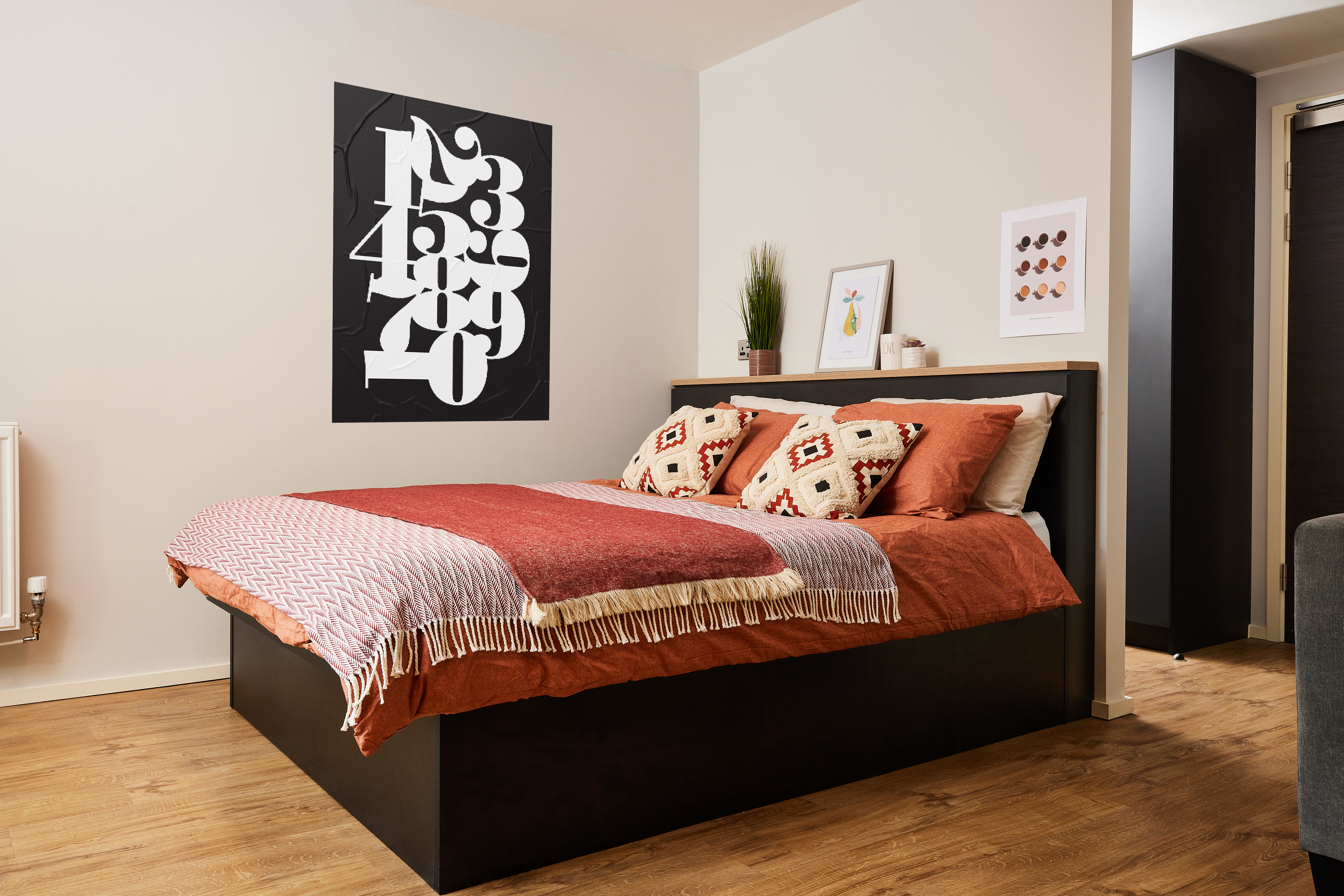 A modern bedroom with a black platform bed featuring red and orange bedding and patterned pillows. A black and white numbered artwork is on the wall. The room has wooden flooring, a small plant, and framed art on a nightstand.