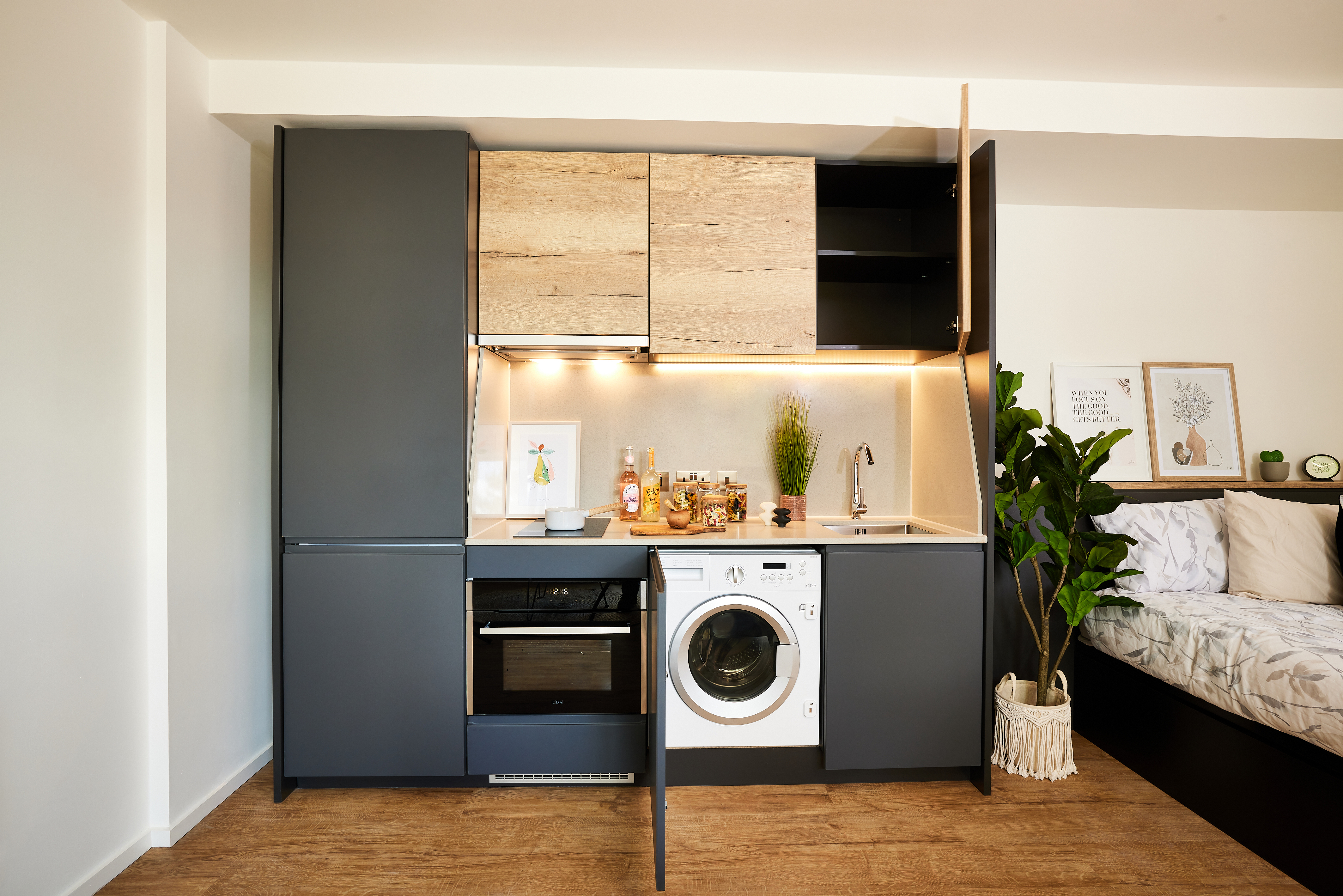 Compact modern kitchenette with black and wood cabinets, an integrated oven, and a washing machine. Bright lighting illuminates the workspace. A bed is partially visible on the right, beneath two Copper Towers artworks. A large leafy plant stands nearby, adding a touch of nature.