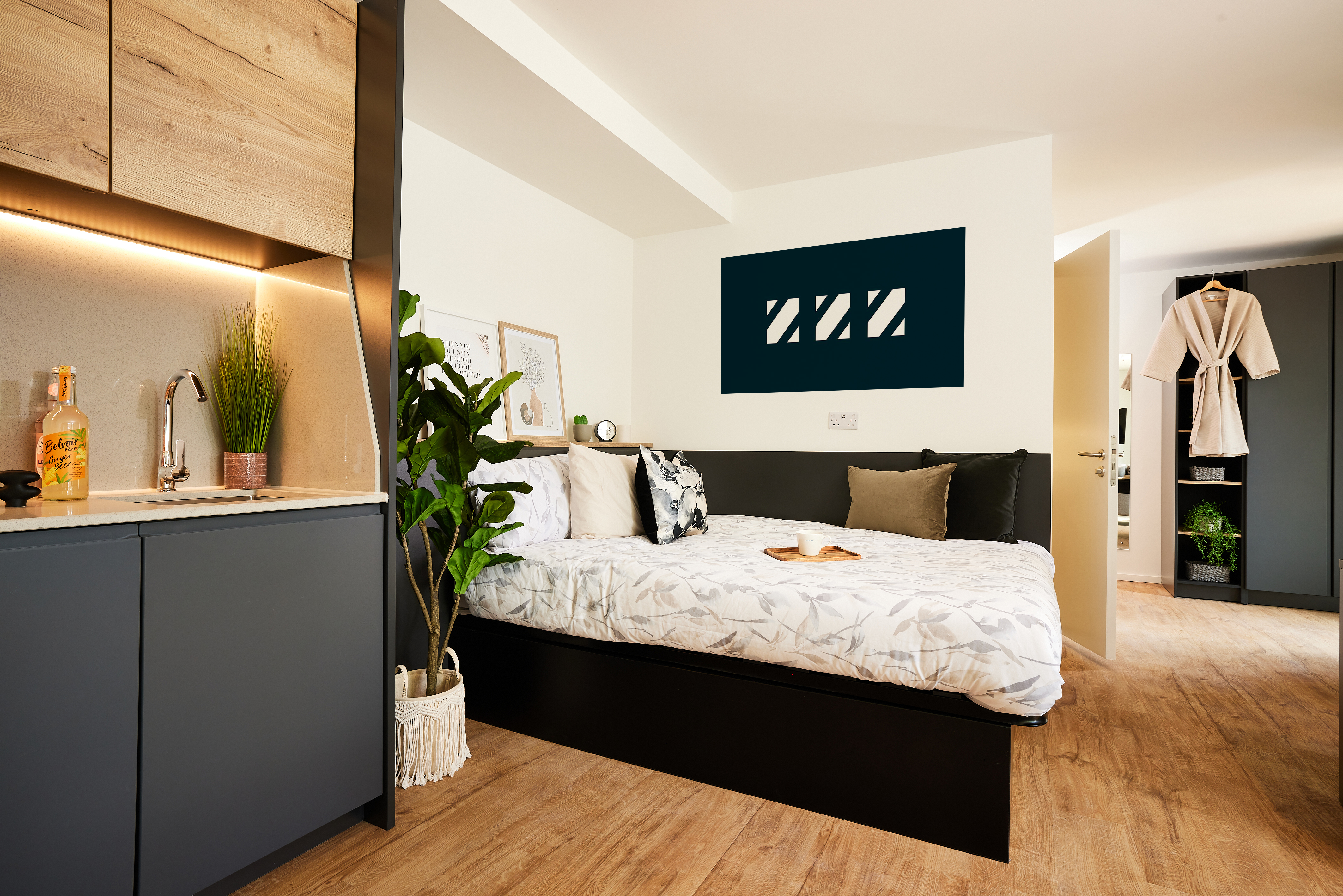 A modern bedroom in Copper Towers showcases a neatly made bed with floral bedding, a wall-mounted abstract art piece, and a potted plant. Adjacent is a kitchenette with wooden cabinets. A wardrobe and hanging robe are visible in the background. The floor is wooden.