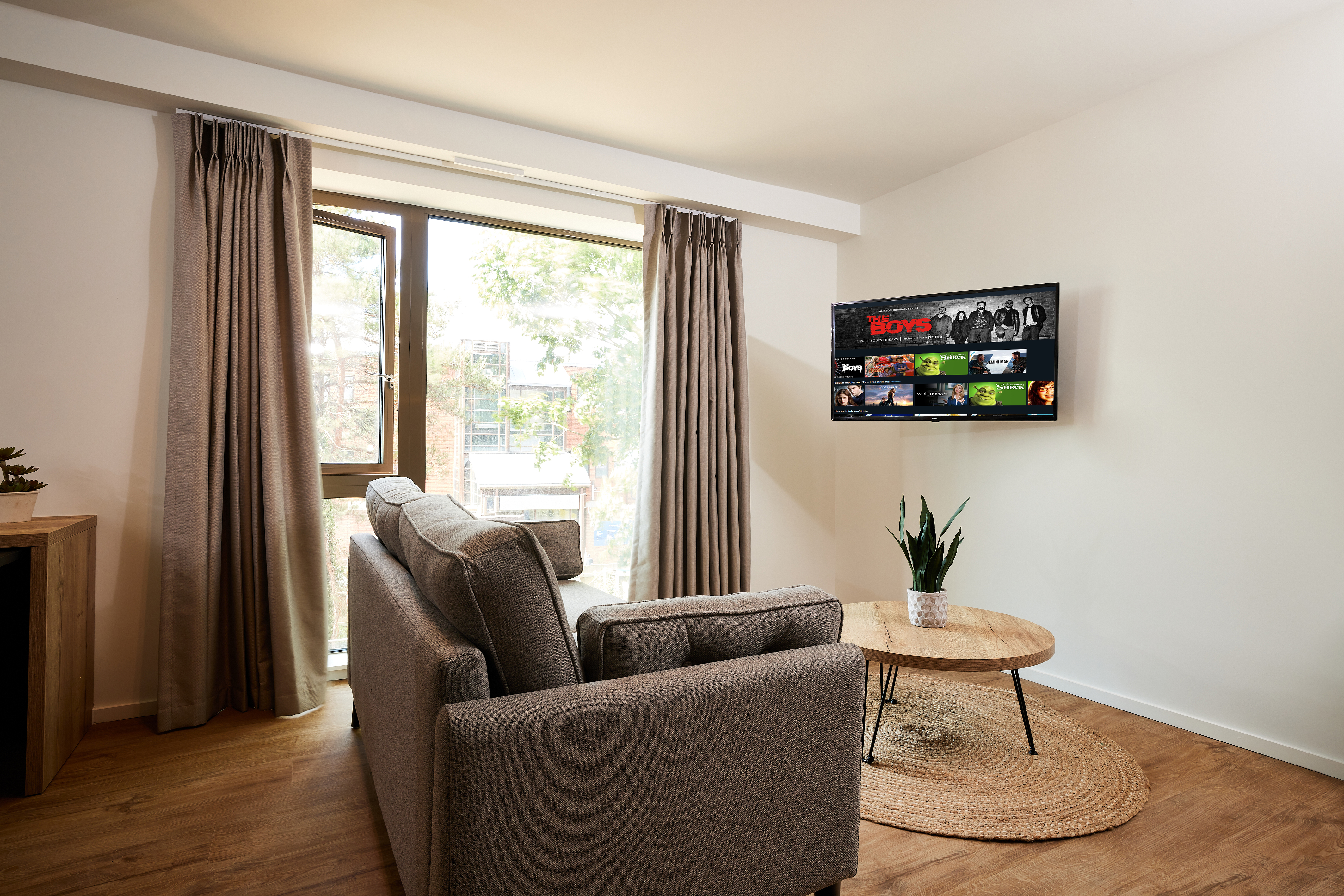 A cozy living room features a gray sofa facing a wall-mounted flat-screen TV showcasing a streaming service interface. A round wooden coffee table with a potted plant rests on a circular rug, while large windows with curtains offer views of the stunning copper architecture outside.